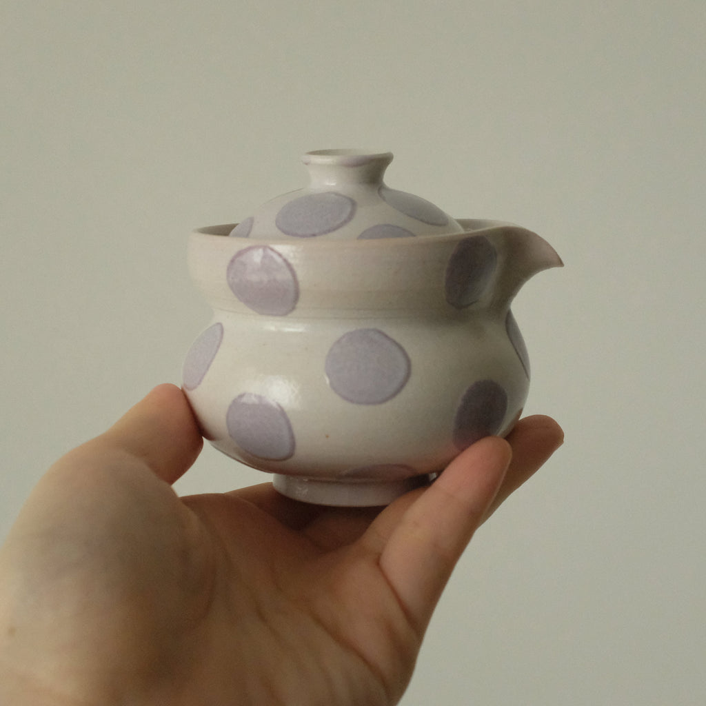 A hand holding a small ceramic teapot with a white glaze and large lavender-purple polka dots, set against a plain light gray background.