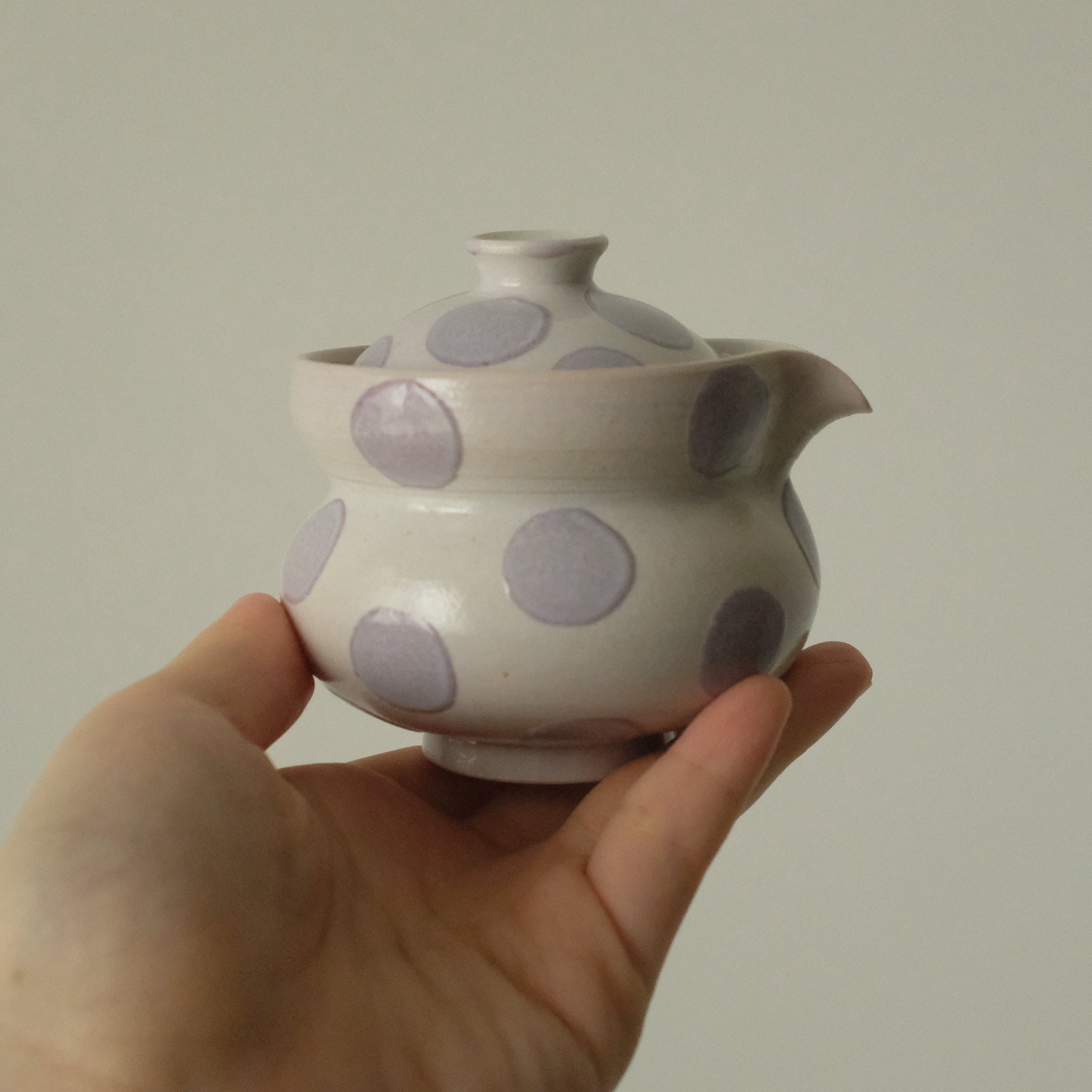 A hand holding a small ceramic teapot with a white glaze and large lavender-purple polka dots, set against a plain light gray background.