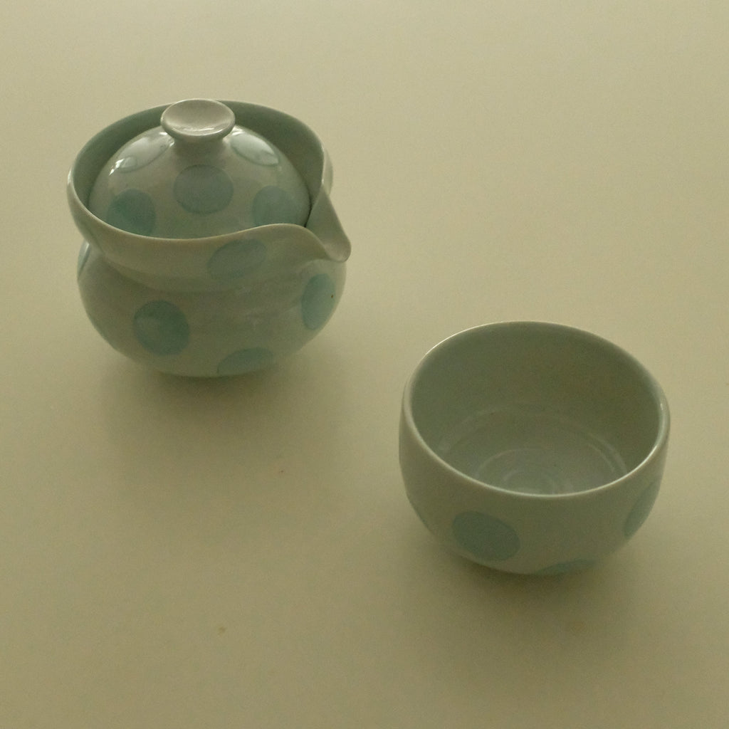 A small ceramic teapot with a matching lid and a small matching cup, both decorated with light blue polka dots on a pale greenish-graice blue glaze, set against a plain beige background.