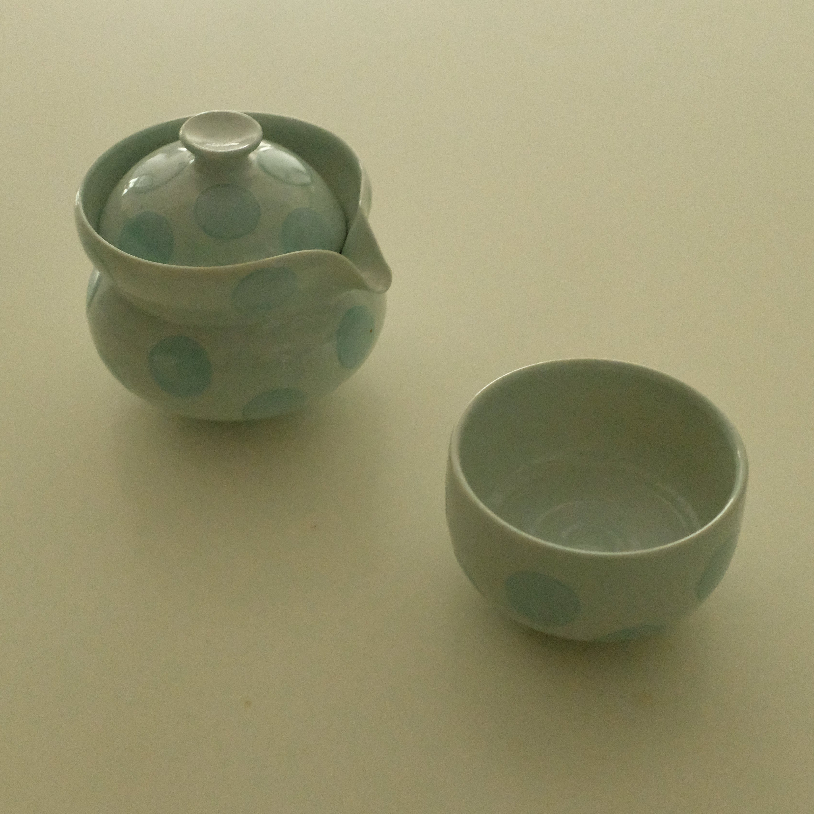 A small ceramic teapot with a matching lid and a small matching cup, both decorated with light blue polka dots on a pale greenish-graice blue glaze, set against a plain beige background.