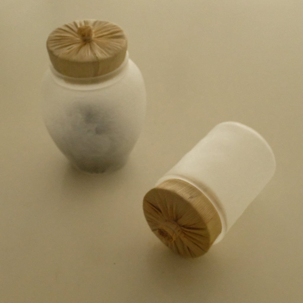 Two frosted glass jars with light wood lids, one standing upright and the other lying on its side, on a plain beige background.