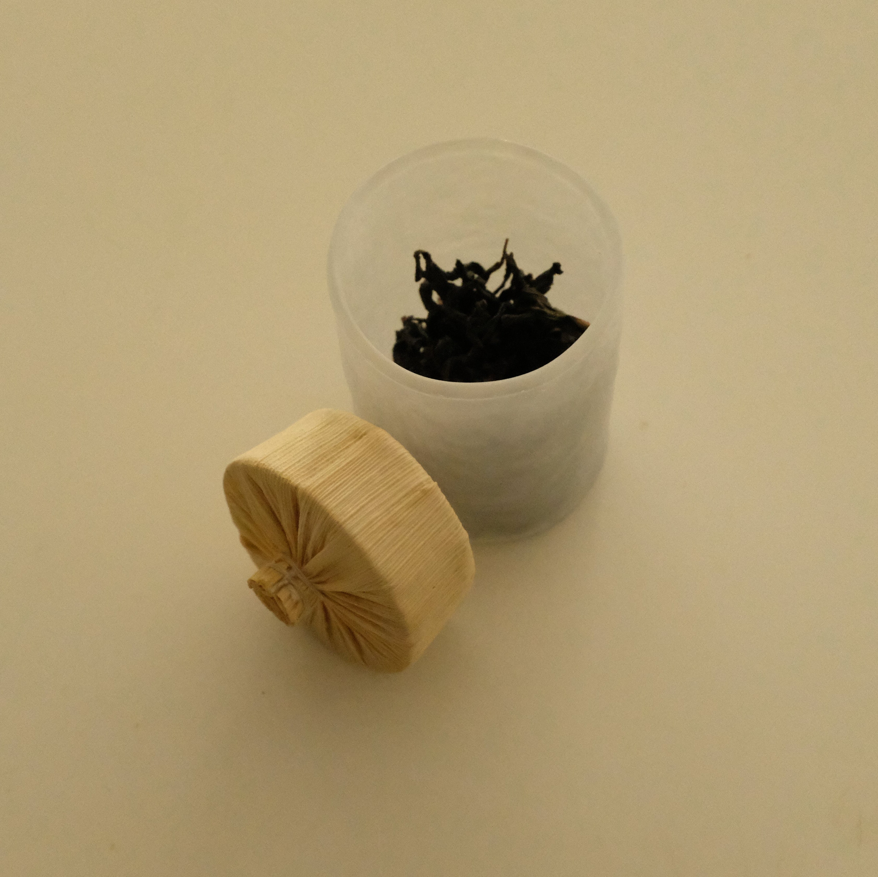 A small clear plastic cup containing dark dried tea leaves, with a light natural wood tamper resting beside it on a plain beige background.