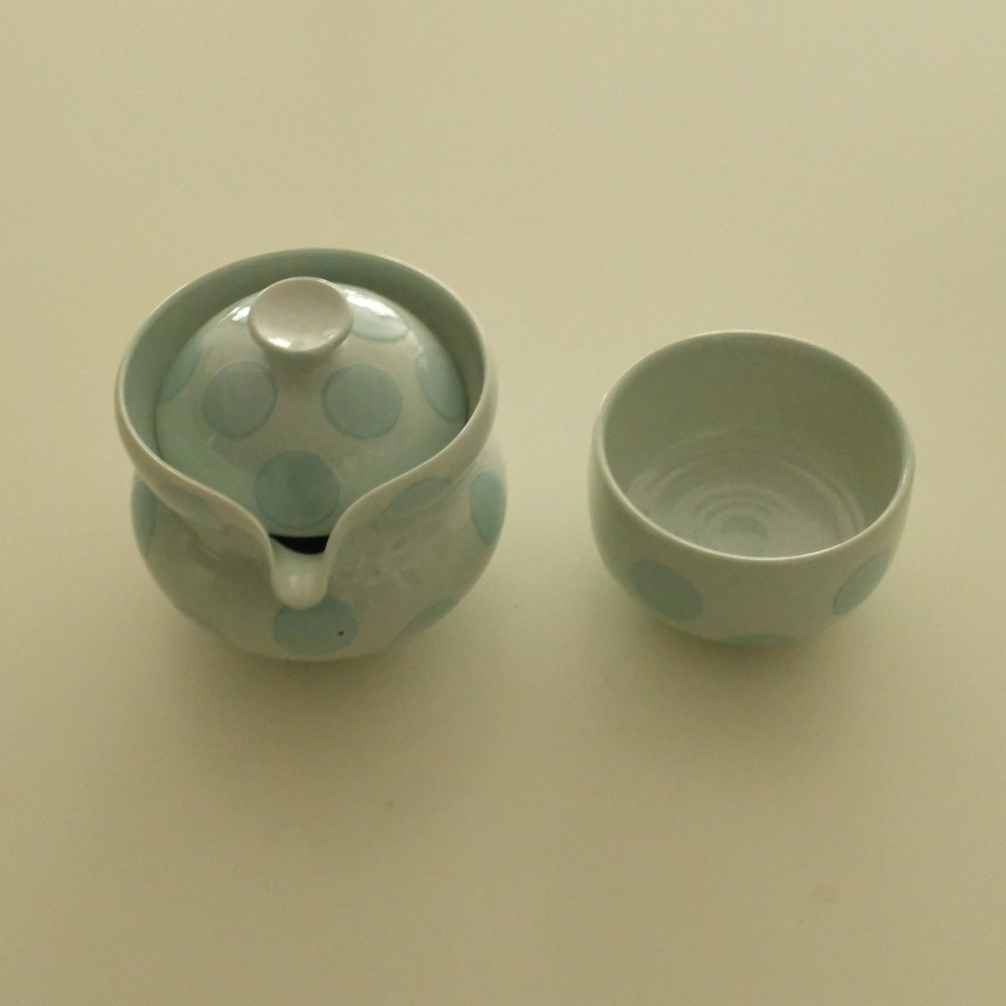 A ceramic handless teapot with a matching small cup, both decorated with a light blue and white polka dot pattern, set against a plain beige background.