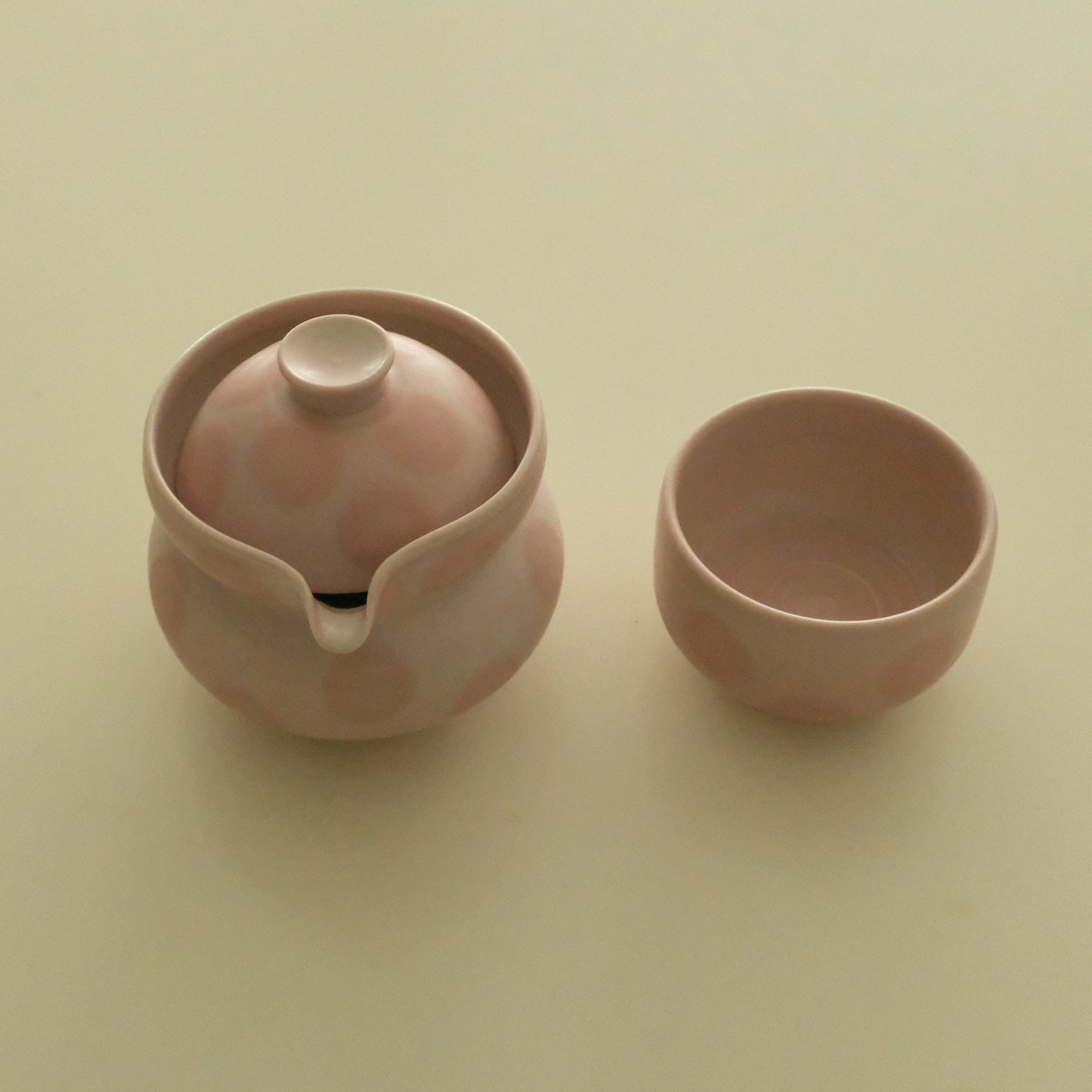 A small pink ceramic teapot with a matching lid and a separate small cup, shown on a plain light beige background.