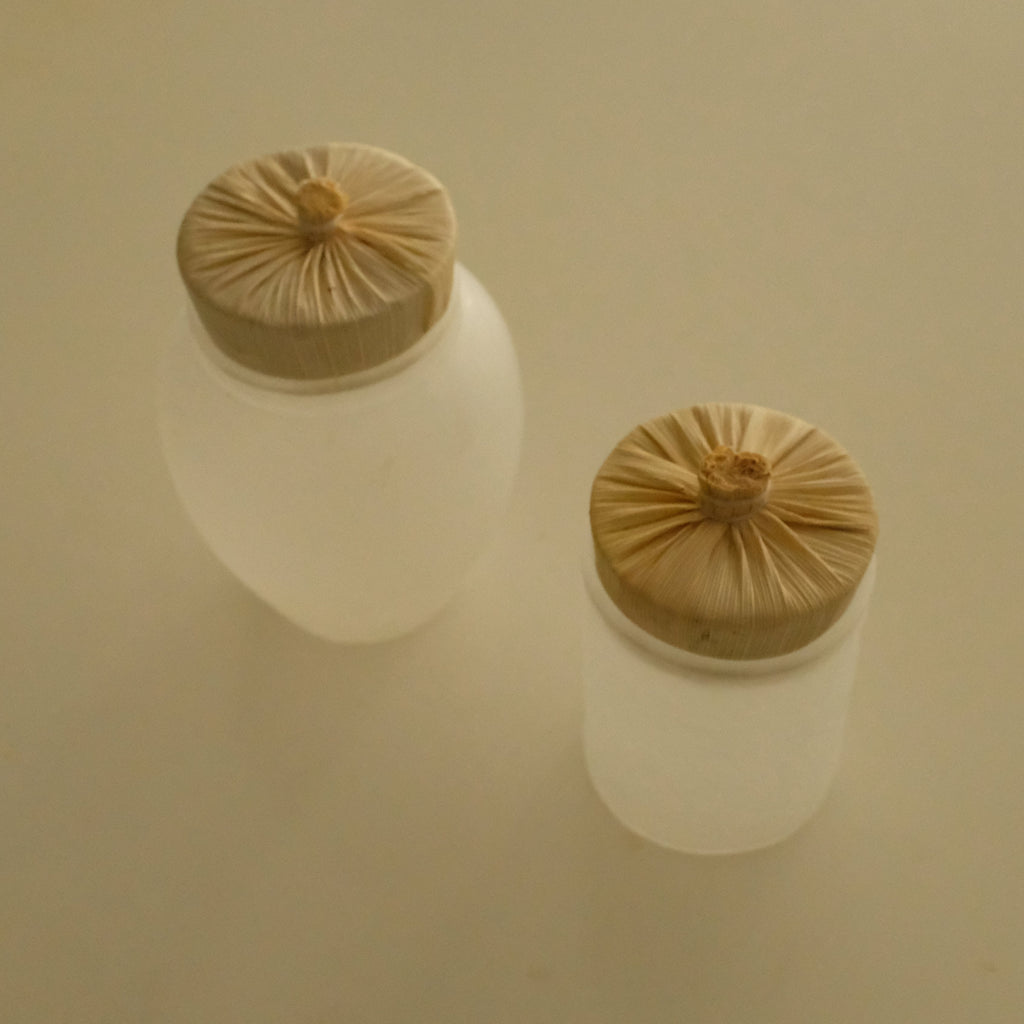 Two frosted glass cosmetic jars with beige fabric-covered screw-top lids, shown on a plain beige background.