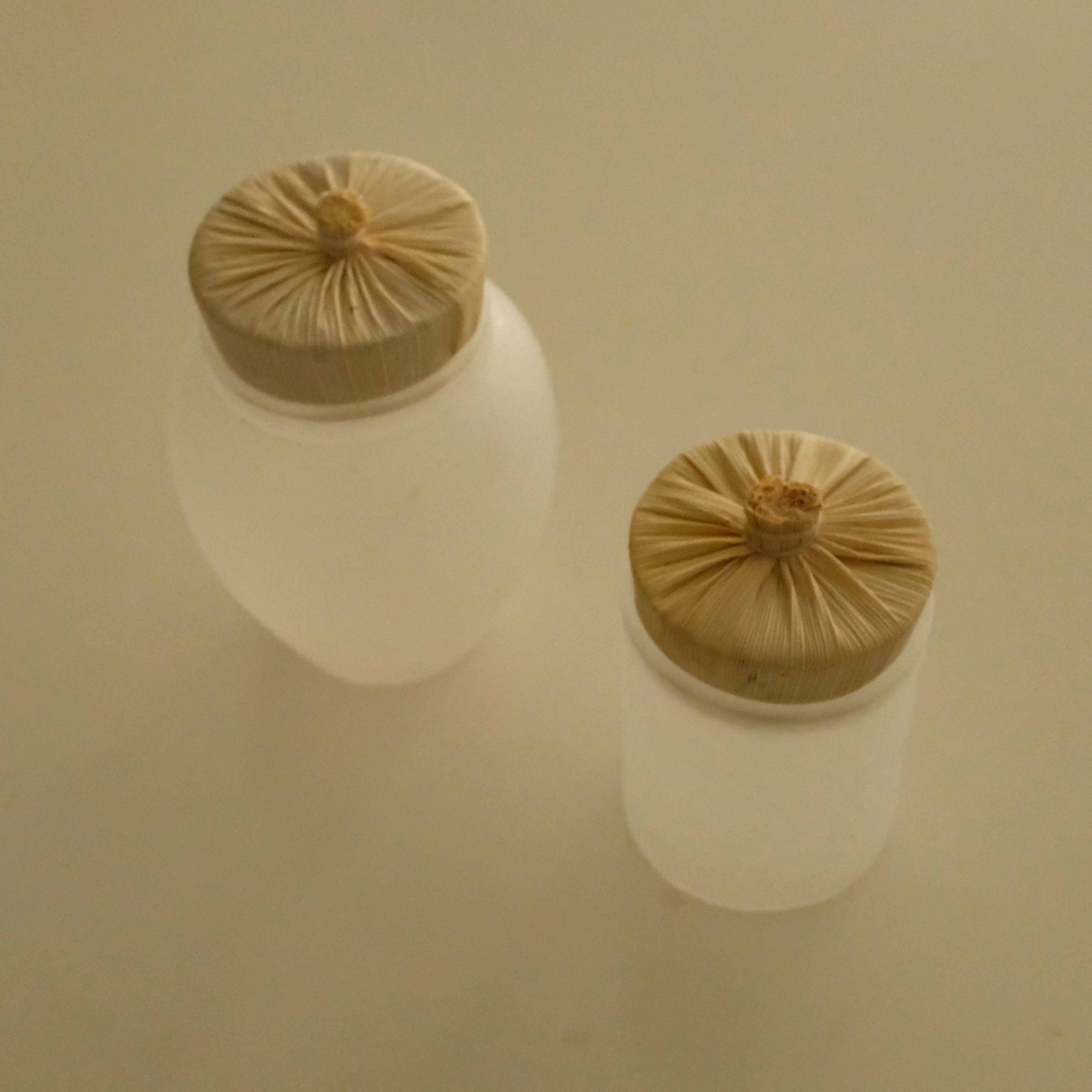 Two frosted glass cosmetic jars with beige fabric-covered screw-top lids, shown on a plain beige background.