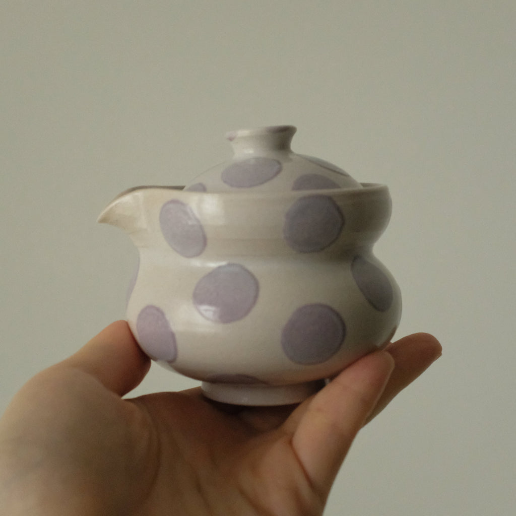 A hand holding a small white ceramic teapot with large lavender-purple polka dots against a plain light gray background.