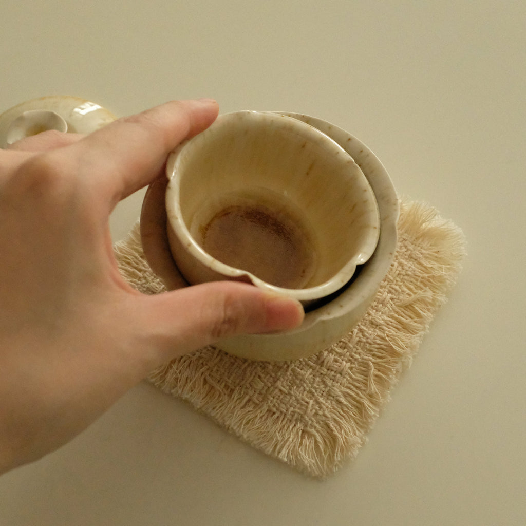 A hand holding a small beige ceramic cup over a textured cream-colored coaster on a plain beige background.