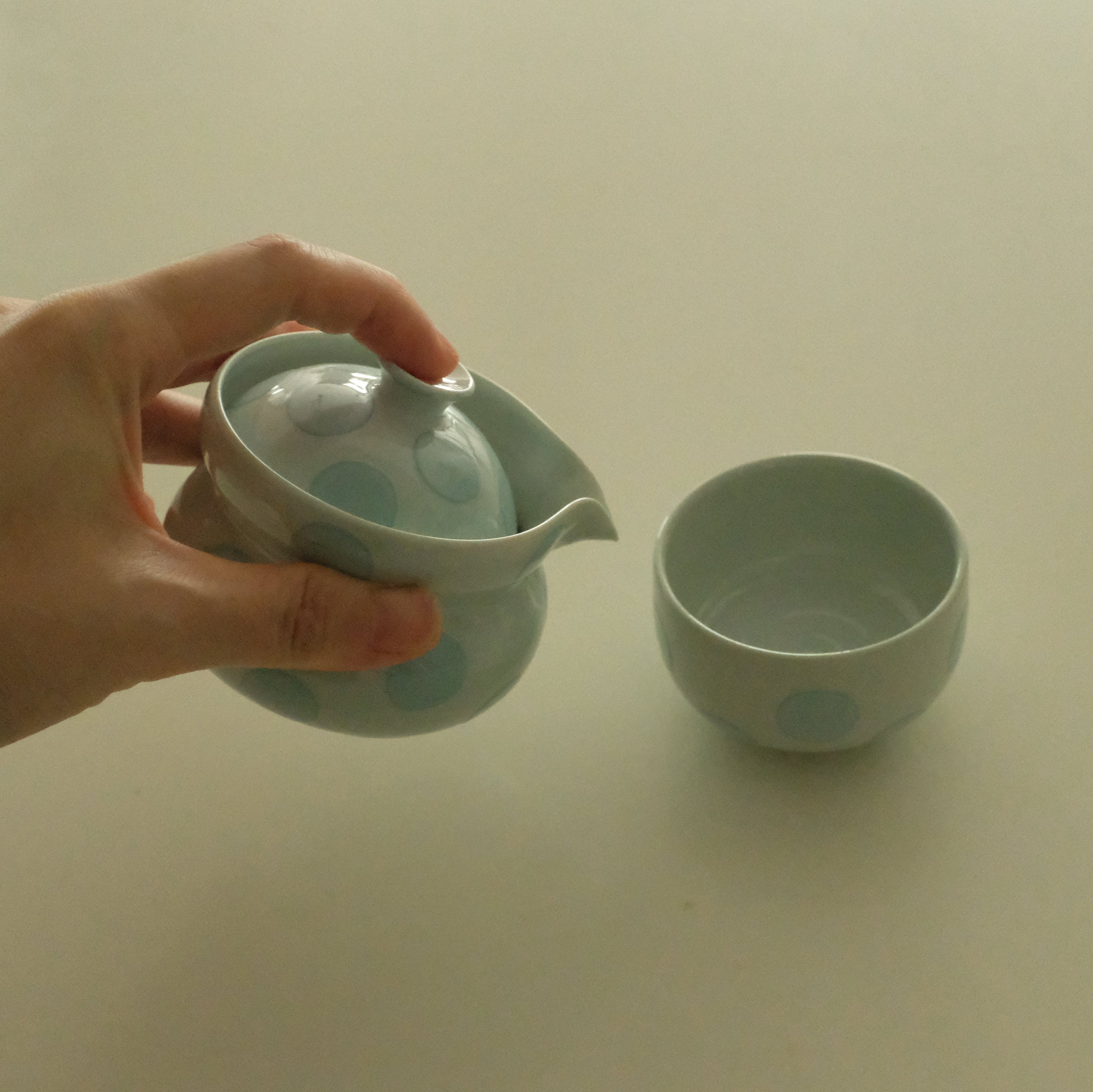 A hand holds a small light blue ceramic teapot with a lid and spout next to a matching light blue ceramic cup on a plain off-white surface.