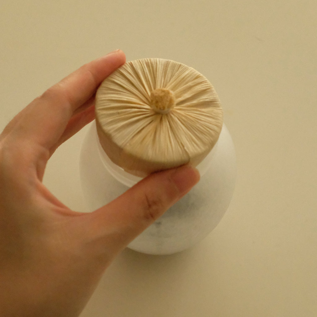 A hand holding the lid of a white jar, which features a round beige fabric saucer with radiating stitched lines and a small wooden knob in the center, set against a plain beige background.