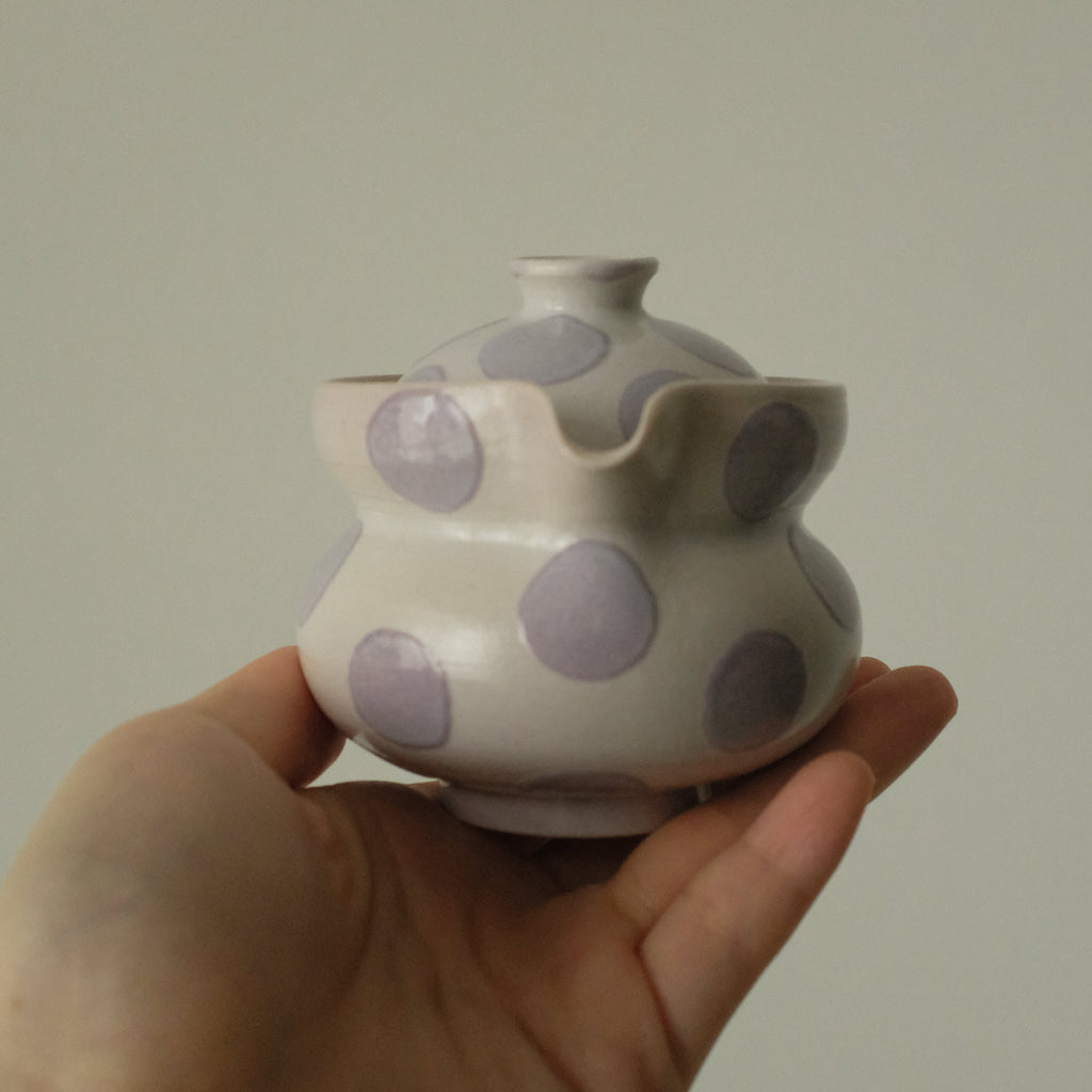 A hand holding a small ceramic teapot with a white glaze and large lavender-purple polka dots, set against a plain light gray background.