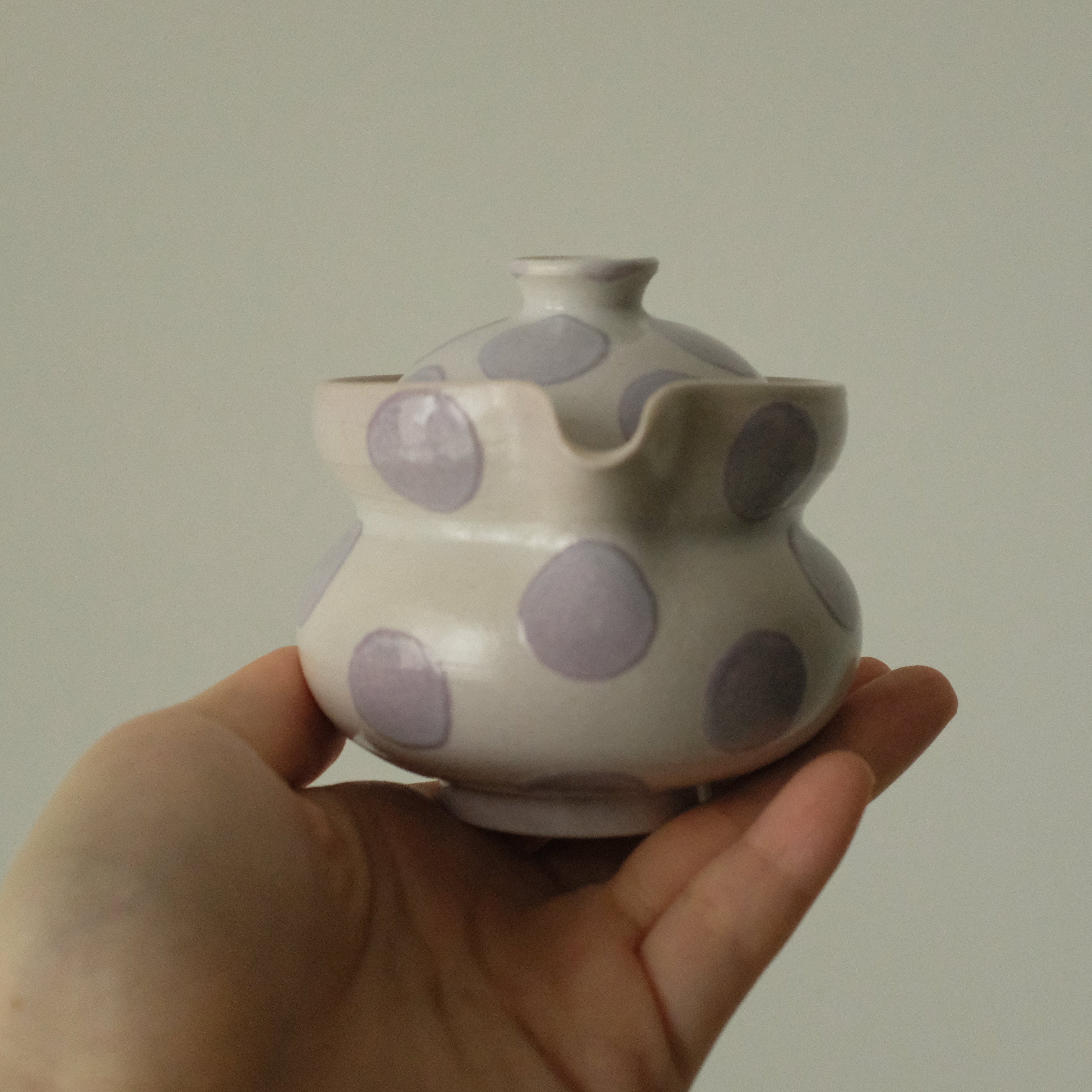 A hand holding a small ceramic teapot with a white glaze and large lavender-purple polka dots, set against a plain light gray background.