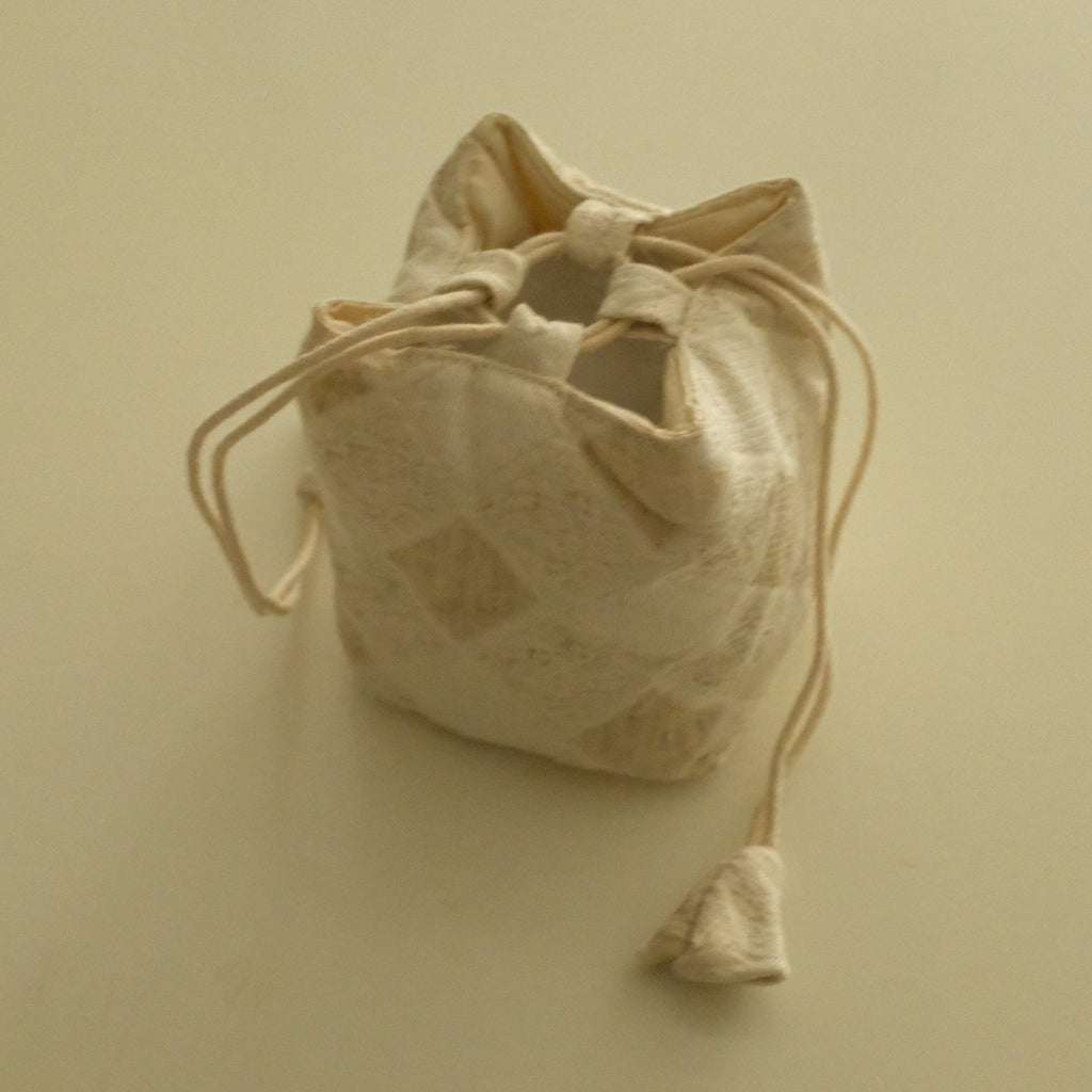A small beige fabric drawstring pouch with a subtle diamond pattern, shown on a plain cream-colored background.