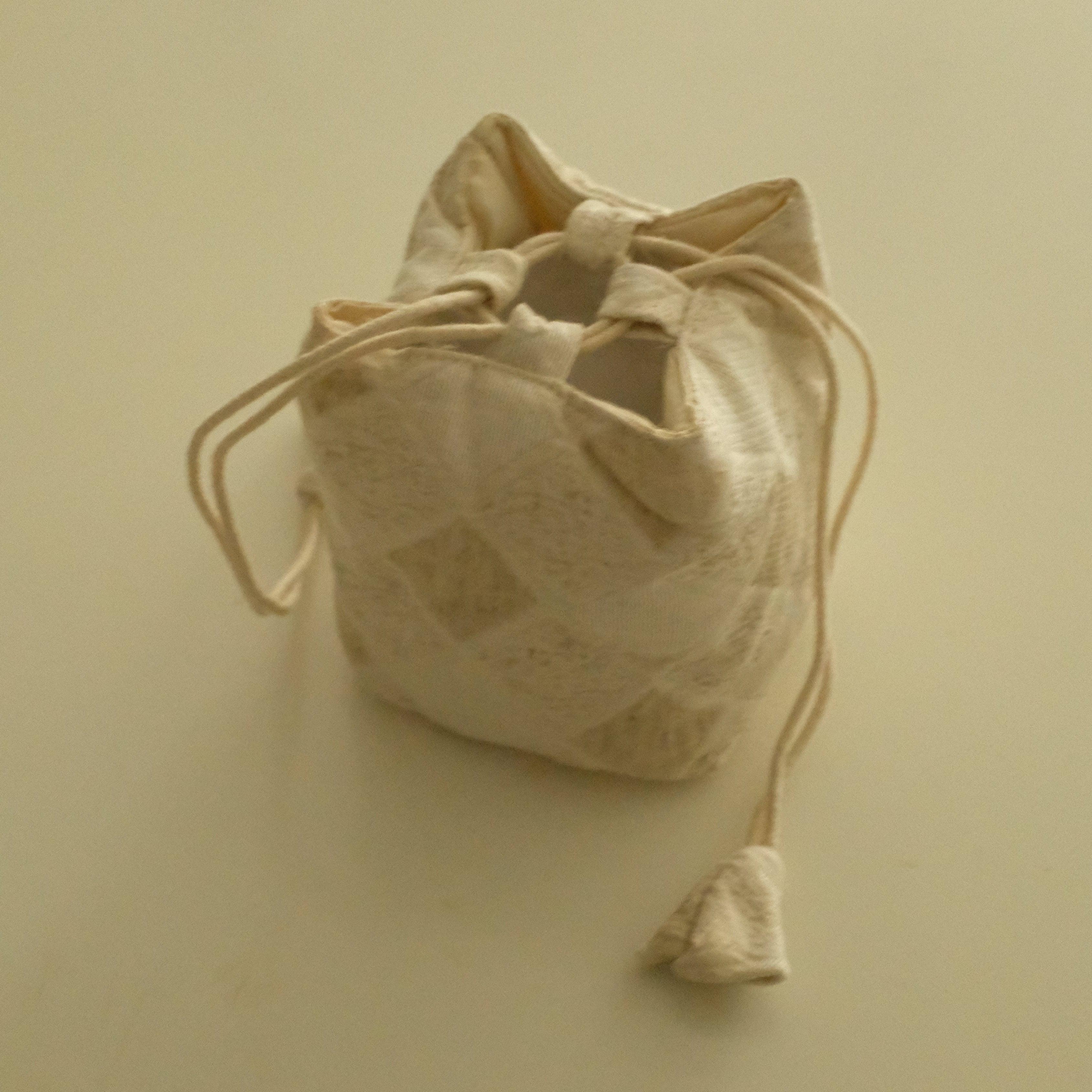 A small beige fabric drawstring pouch with a subtle diamond pattern, shown on a plain cream-colored background.