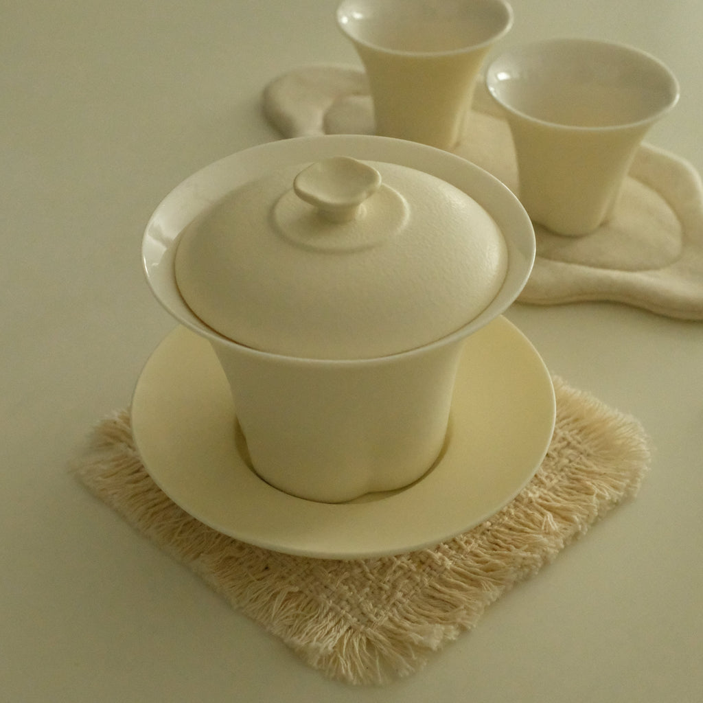 A cream-colored ceramic tea set, featuring a lidded gaiwan on a saucer, with two small cups on a matching cloth coaster, arranged on textured fringed coasters on a light beige surface.
