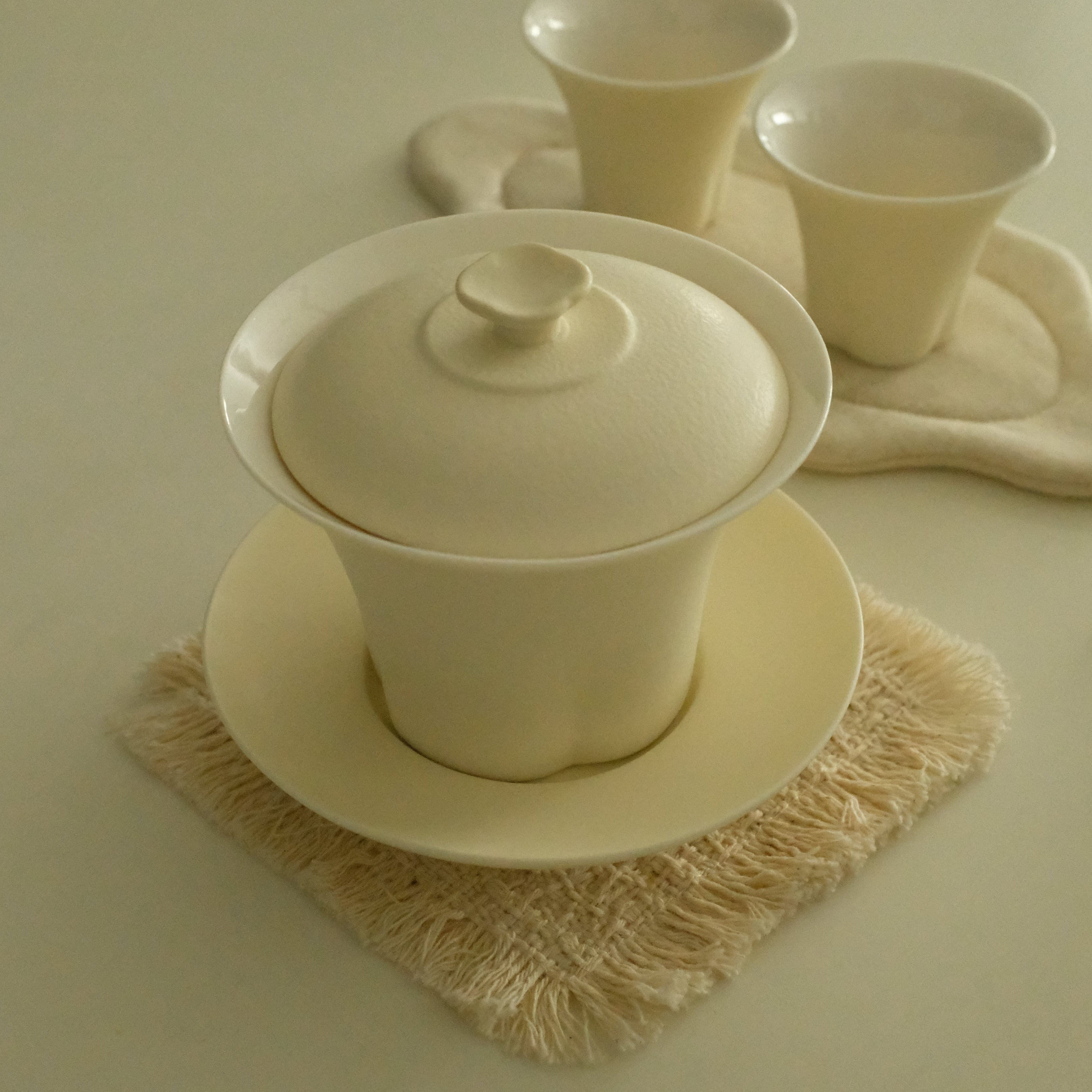 A cream-colored ceramic tea set, featuring a lidded gaiwan on a saucer, with two small cups on a matching cloth coaster, arranged on textured fringed coasters on a light beige surface.
