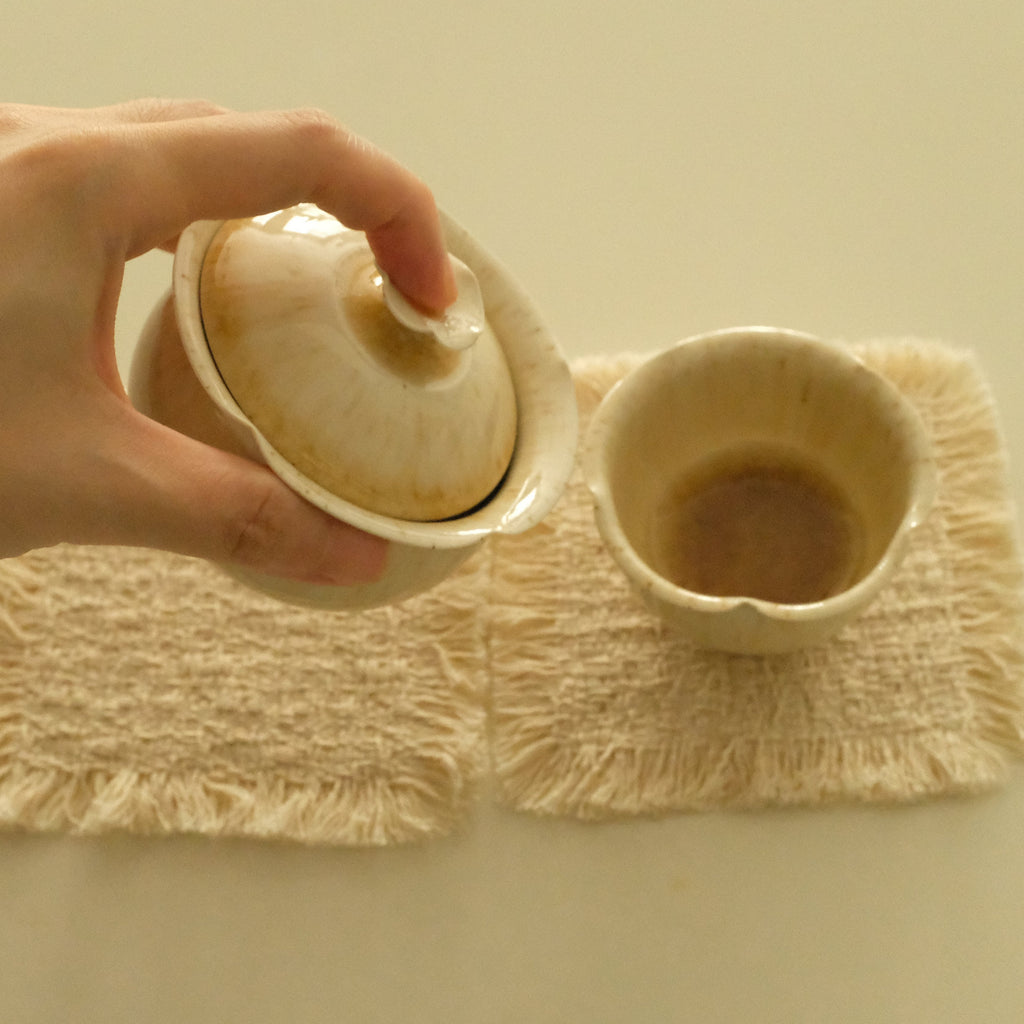 A hand holds a small beige ceramic teapot lid above a matching beige cup resting on two cream woven placemats on a light beige surface.