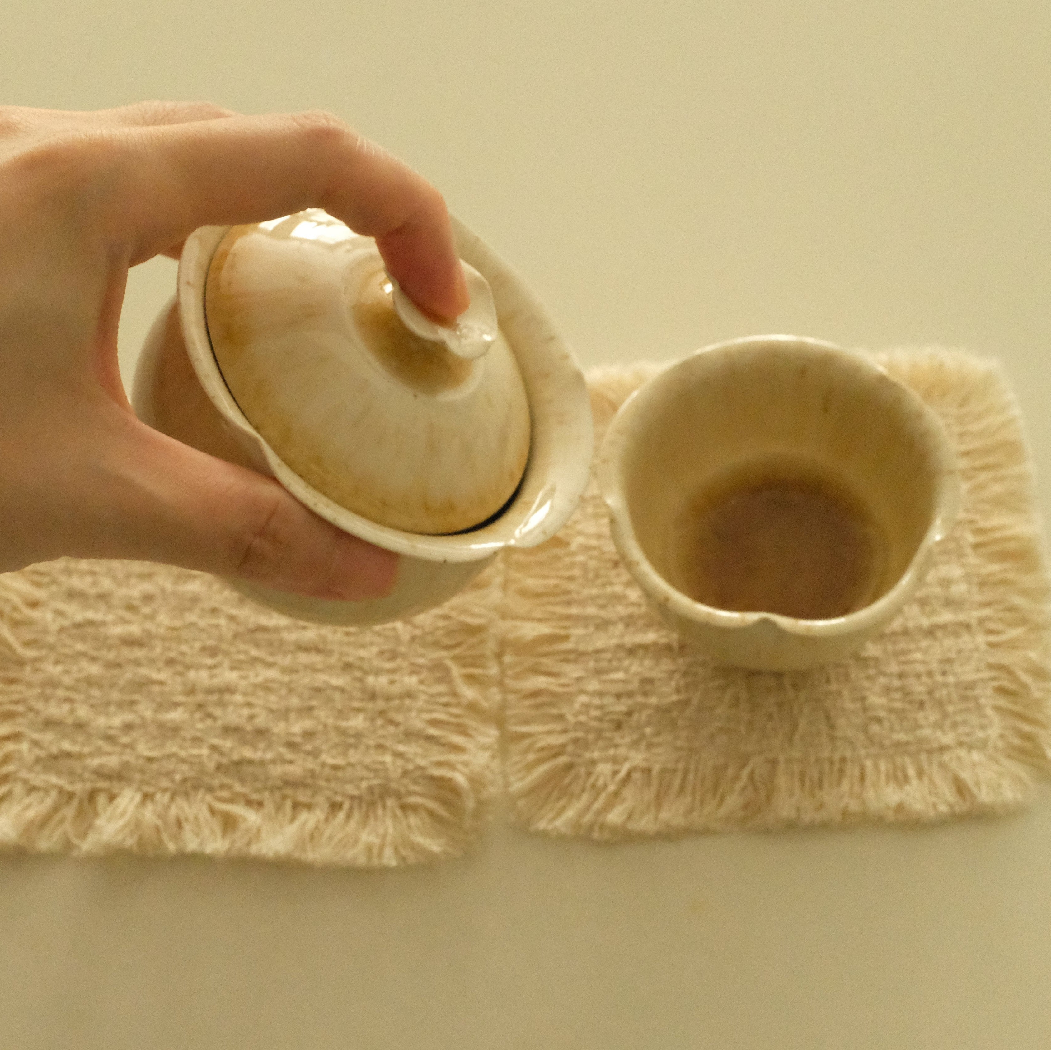 A hand holds a small beige ceramic teapot lid above a matching beige cup resting on two cream woven placemats on a light beige surface.