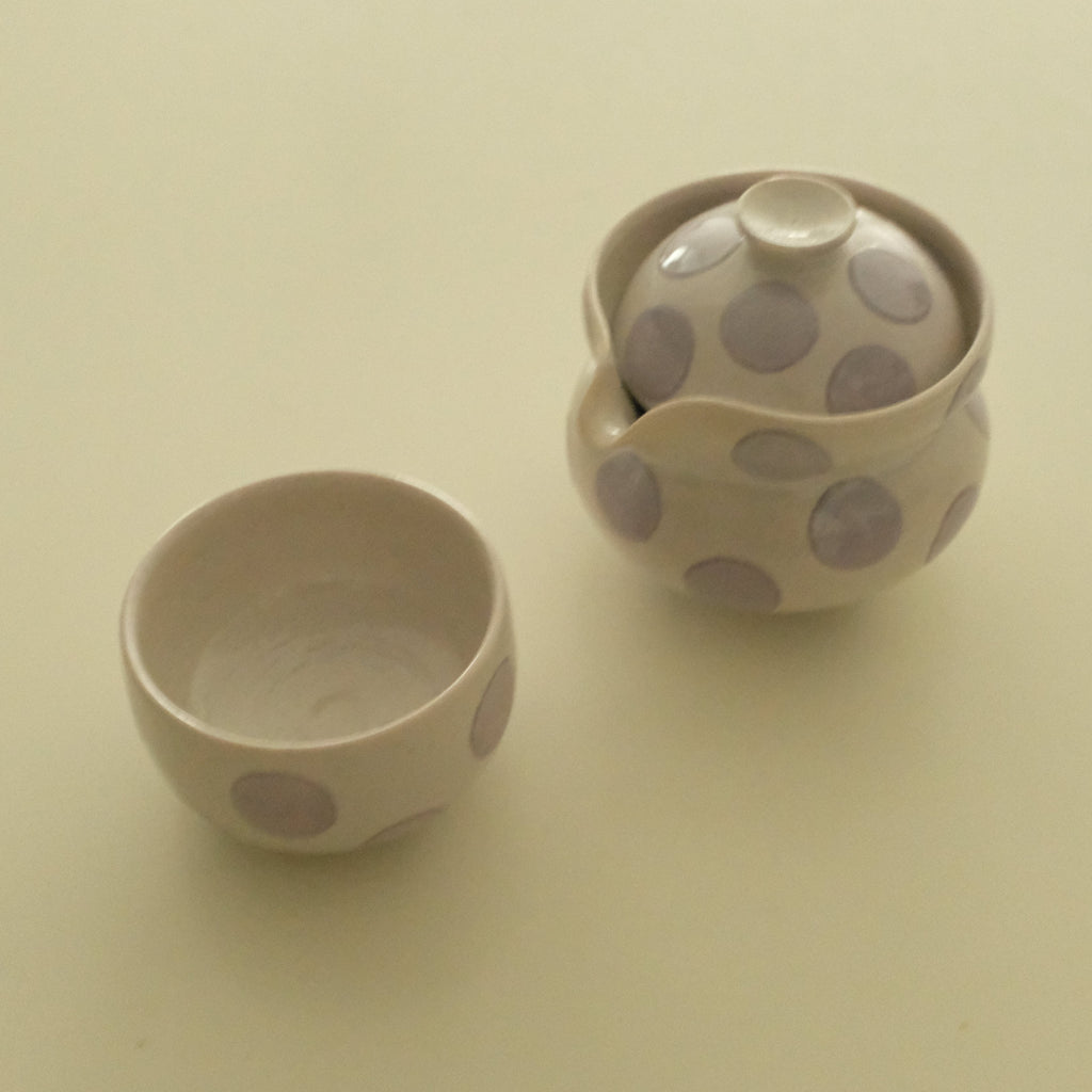 A ceramic tea set with a small lidded teapot and a matching small cup, both decorated with large lavender polka dots on a warm cream background, set against a plain cream backdrop.
