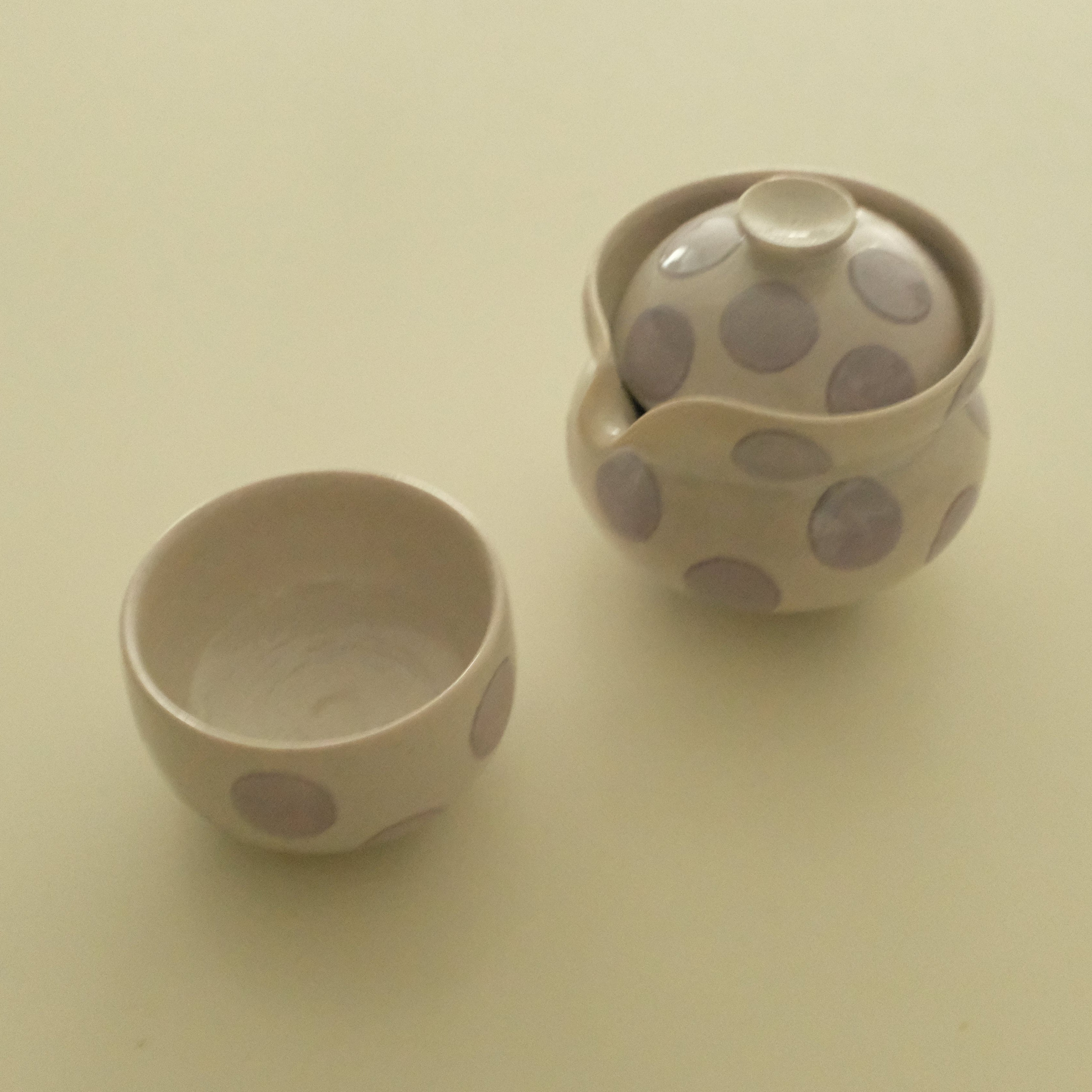 A ceramic tea set with a small lidded teapot and a matching small cup, both decorated with large lavender polka dots on a warm cream background, set against a plain cream backdrop.