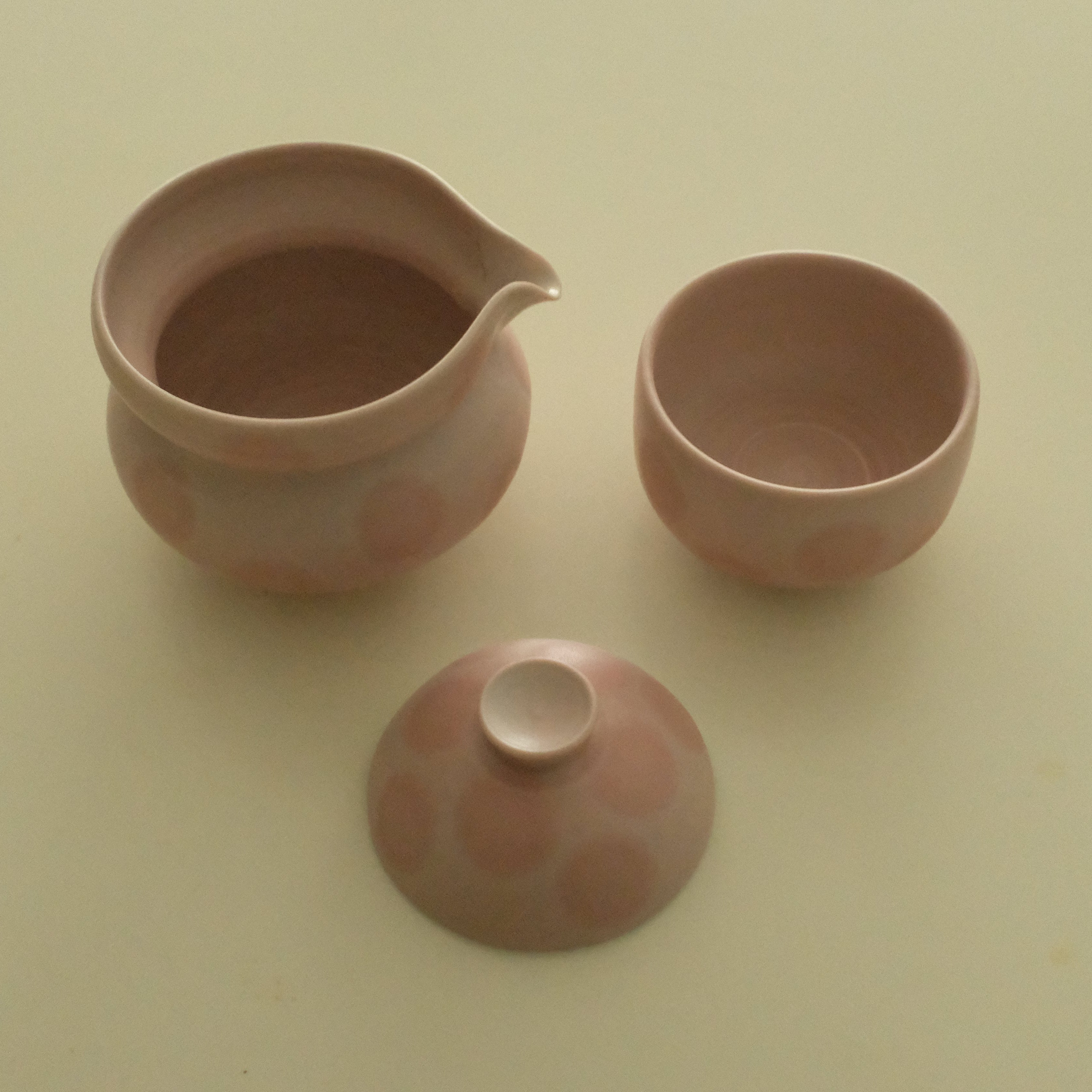 A three-piece ceramic tea set in a dusty pink color, including a teapot, a small cup, and a lid, arranged on a plain light beige background.