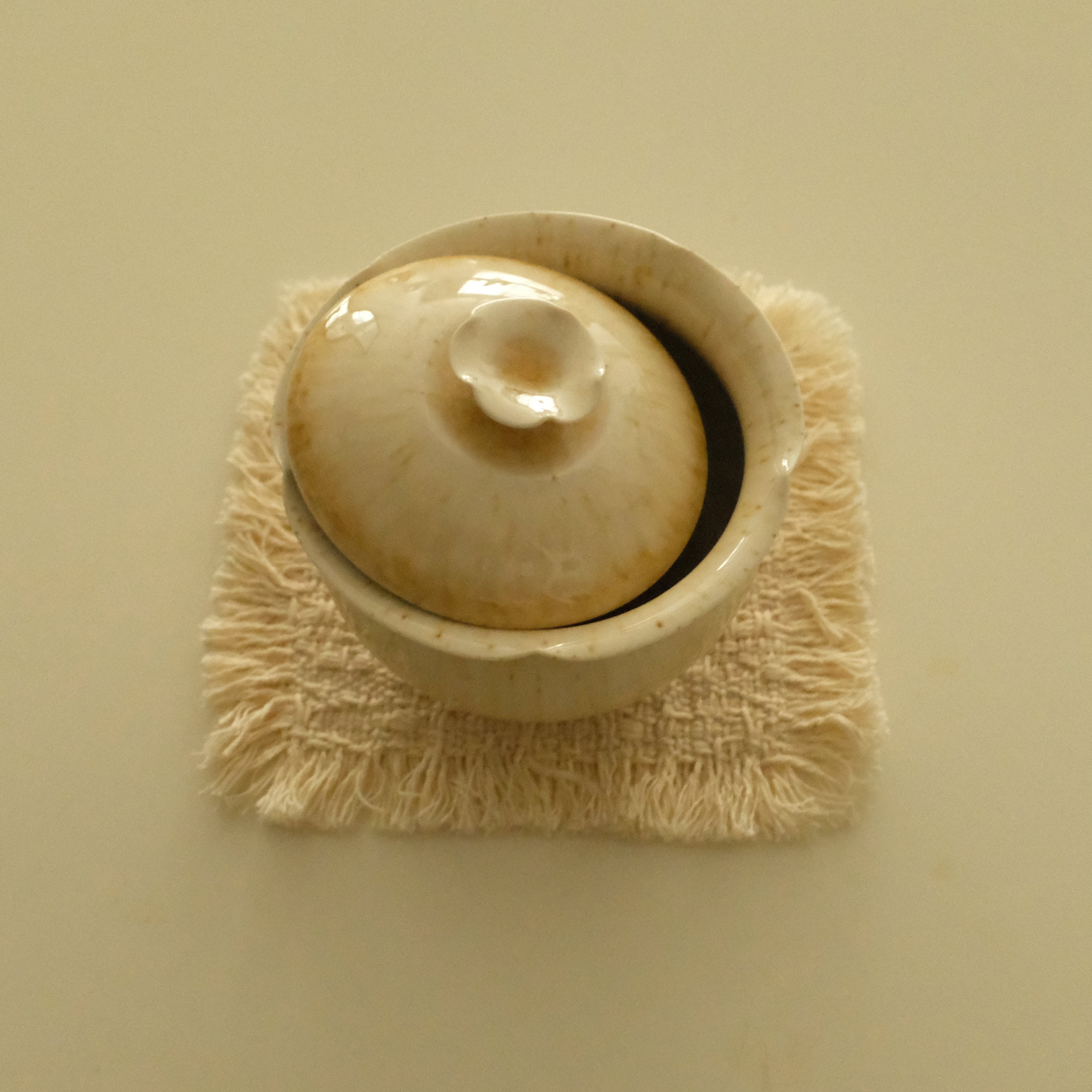 A small beige ceramic lidded bowl or tureen with a textured cream fringed mat underneath, photographed from above on a plain beige background.