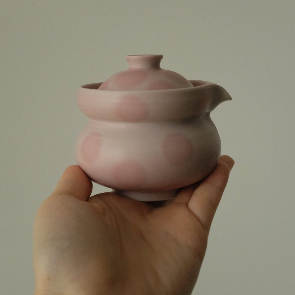 A small pink ceramic teapot with a lid and spout, held in a person's hand against a plain light gray background.