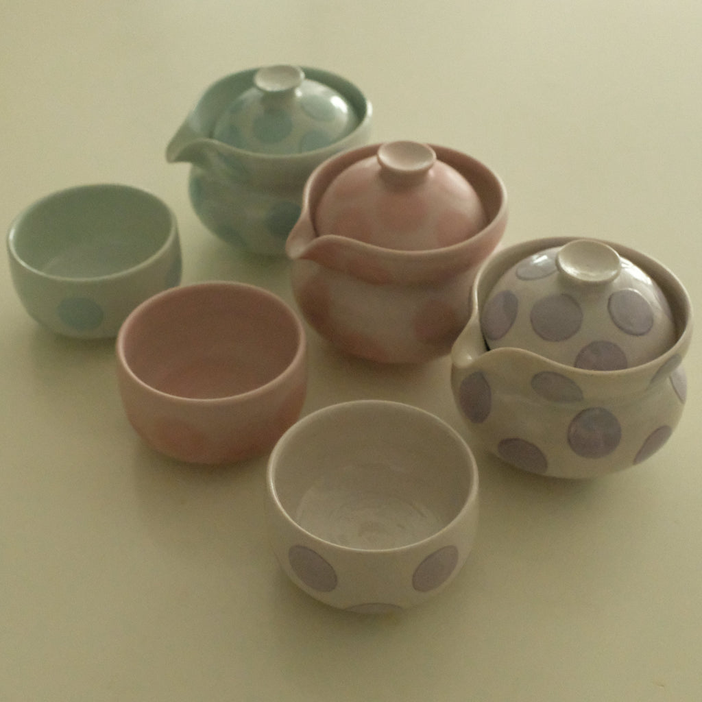 A set of six pastel-colored ceramic tea and tea-time items, including a teapot, two teacups, a small lidded pot, and two bowls, decorated with polka dots in soft pink, lavender, mint green, and cream colors.