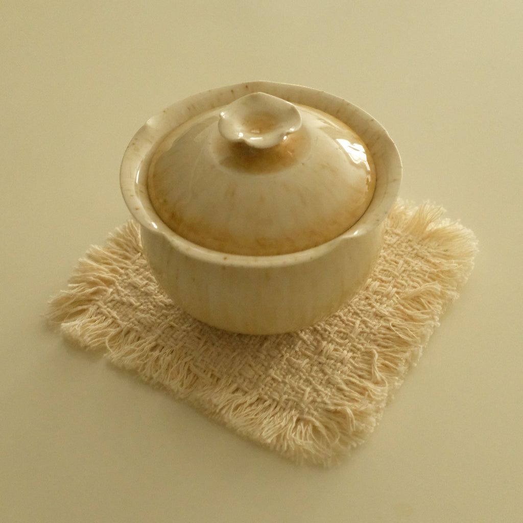 A small beige ceramic lidded bowl or teapot-style vessel sits on a textured cream fringed coaster against a plain warm beige background.
