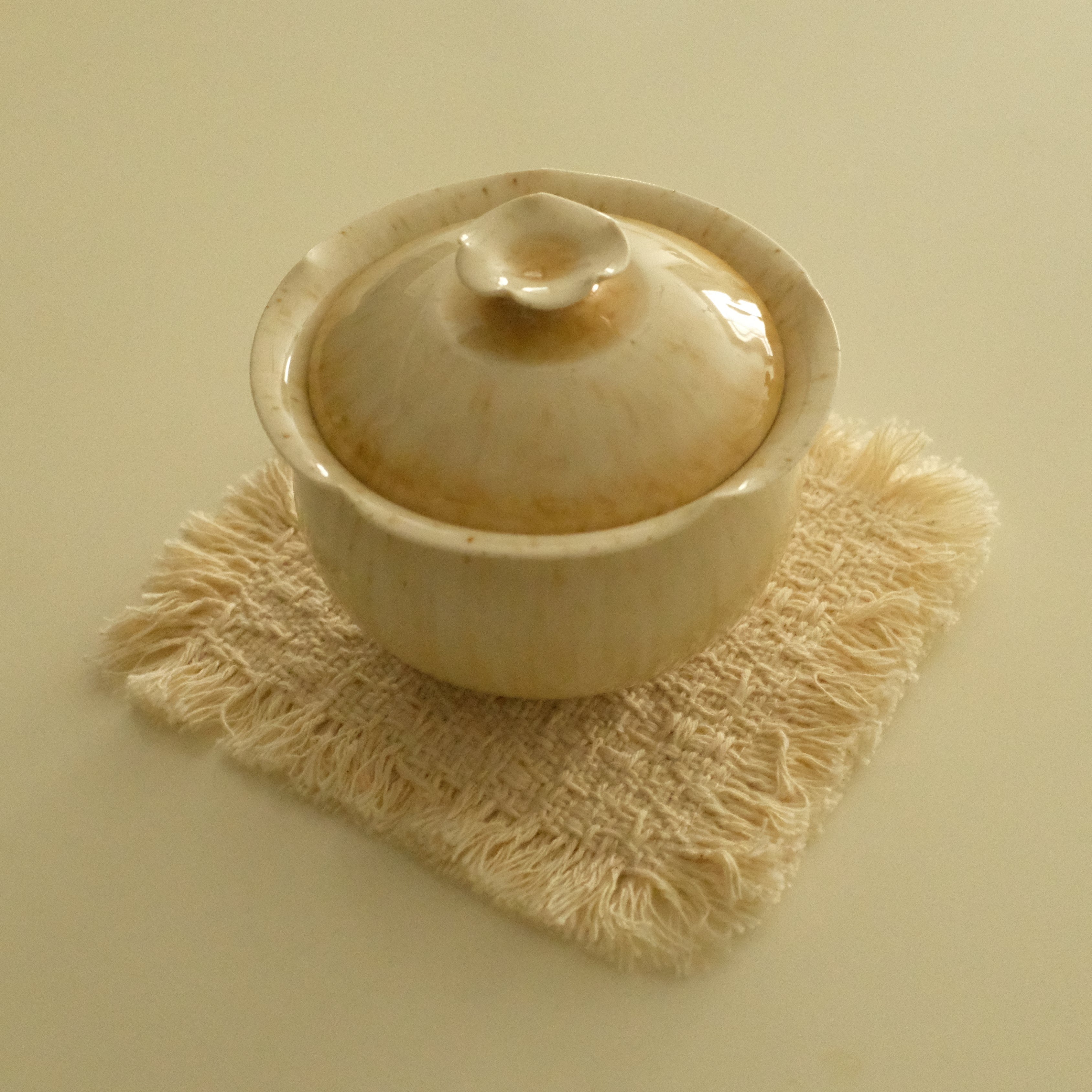 A small beige ceramic lidded bowl or teapot-style vessel sits on a textured cream fringed coaster against a plain warm beige background.