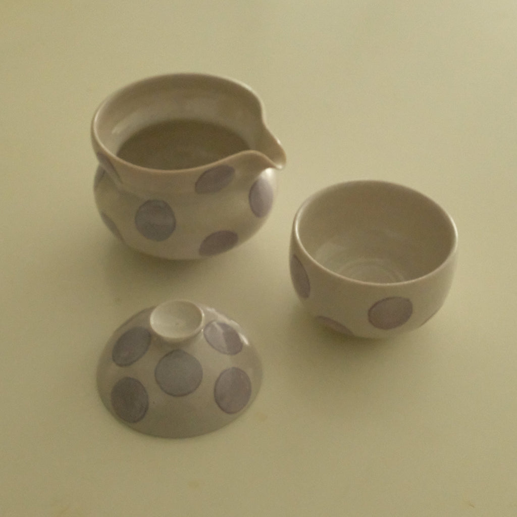 A ceramic tea set with a lid, small handless teapot and cup, decorated with large gray polka dots on a warm cream background, set against a plain cream backdrop.