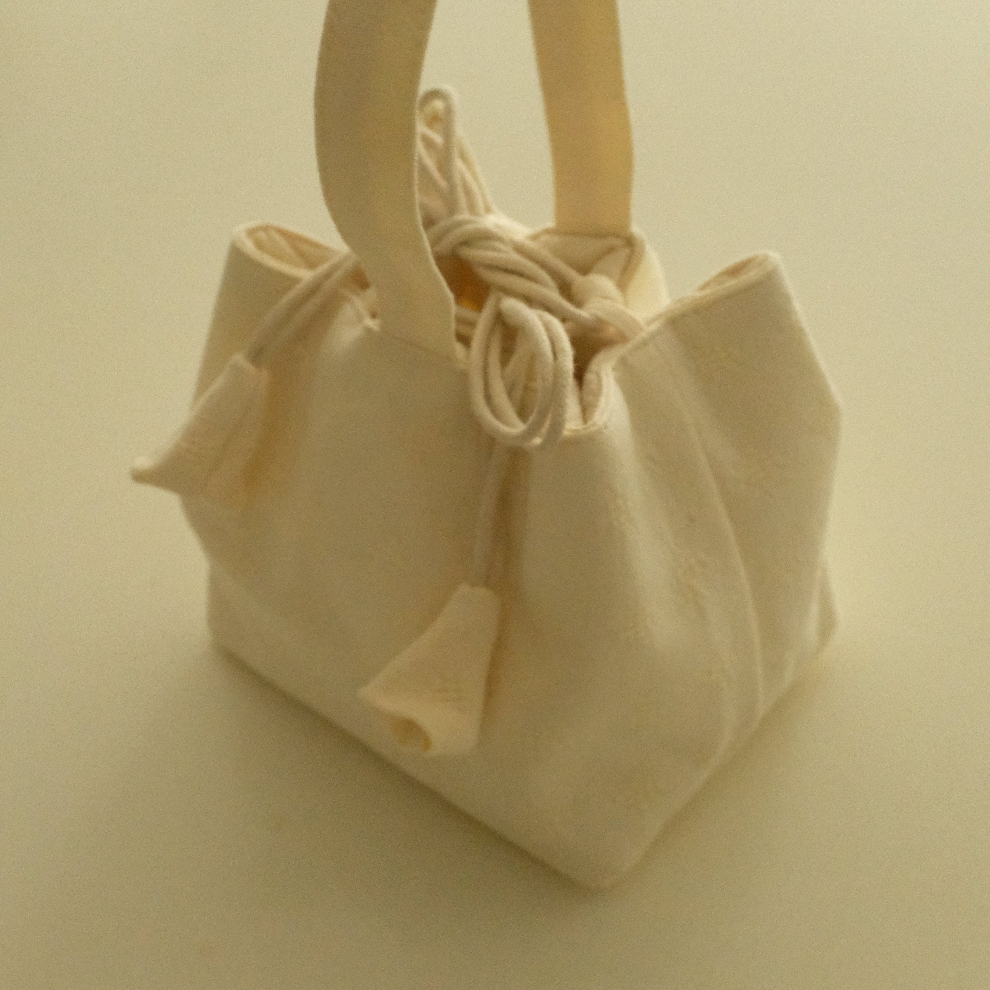 A small cream canvas drawstring bag with two top handles, photographed on a plain warm beige background.