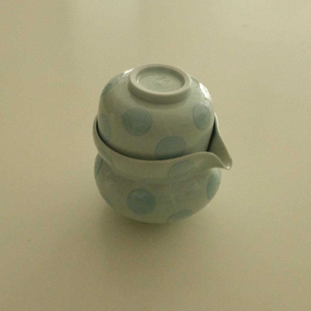 A small ceramic teapot with a matching lid, decorated with light blue polka dots on an off-white glaze, set against a plain beige background.