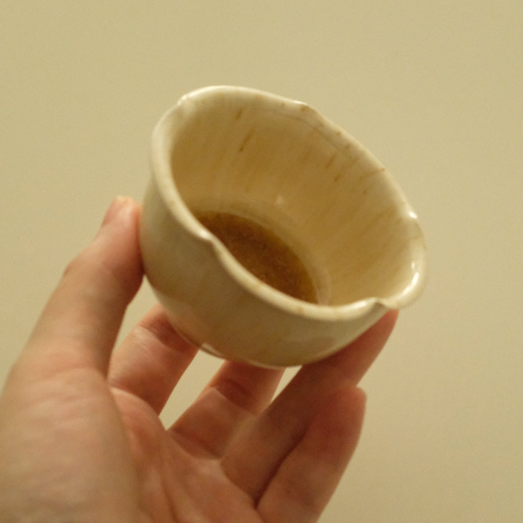 A hand holding a small handmade ceramic cup with a scalloped rim and a warm cream glaze, set against a plain beige background.