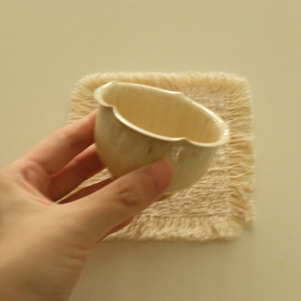 A hand holding a small cream-colored ceramic cup with a scalloped rim, positioned in front of a textured cream fabric square against a solid cream background.