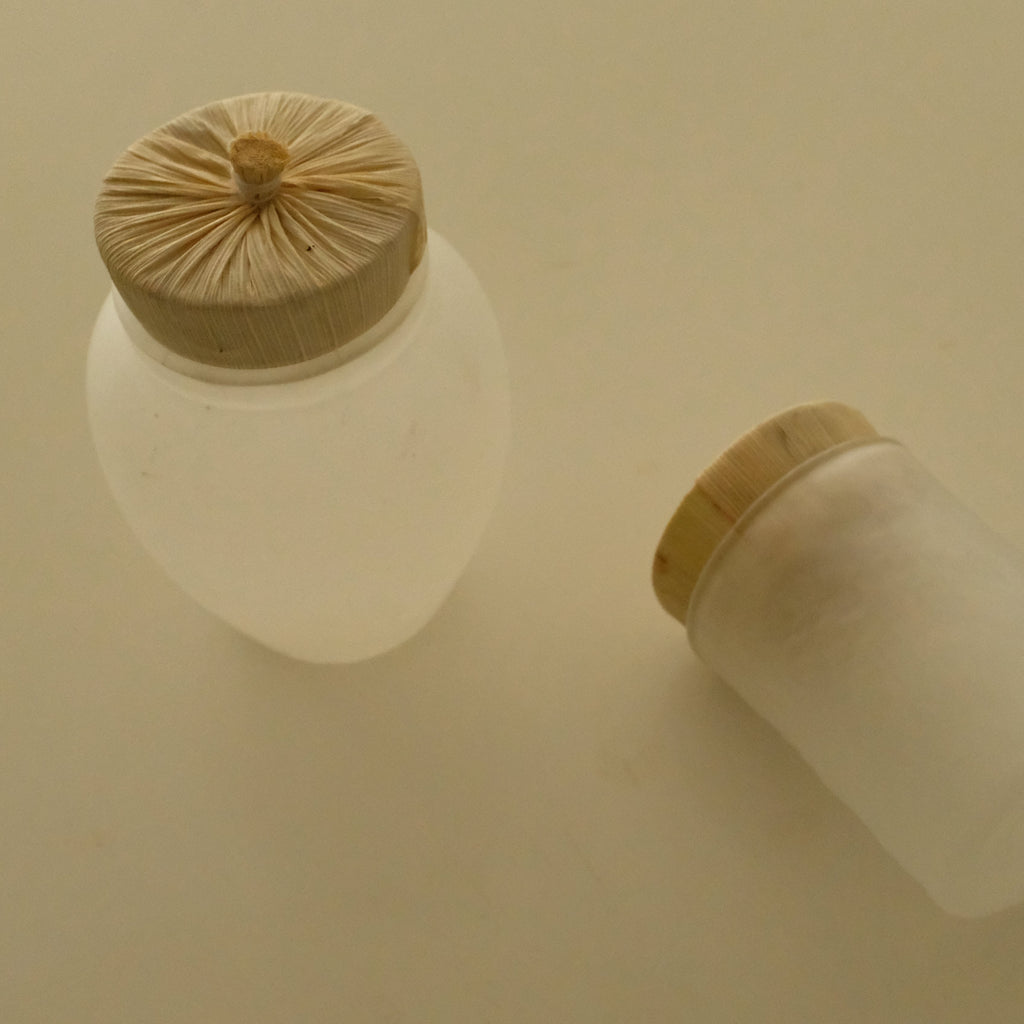 Two frosted glass jars with light wood lids on a warm beige background, one standing upright and the other lying on its side.