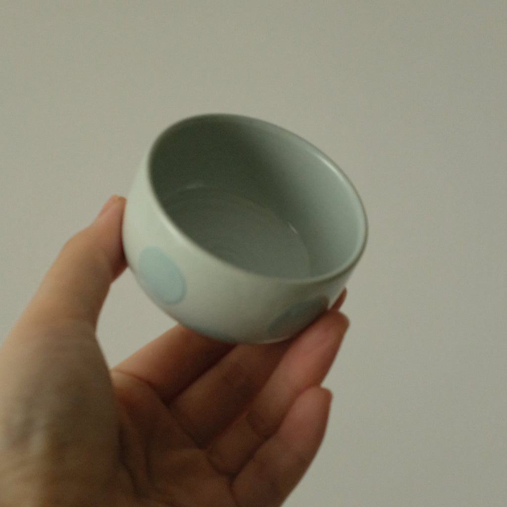 A hand holding a small ceramic cup with a pale blue-green glaze and light blue circular dot patterns, set against a plain light gray background.