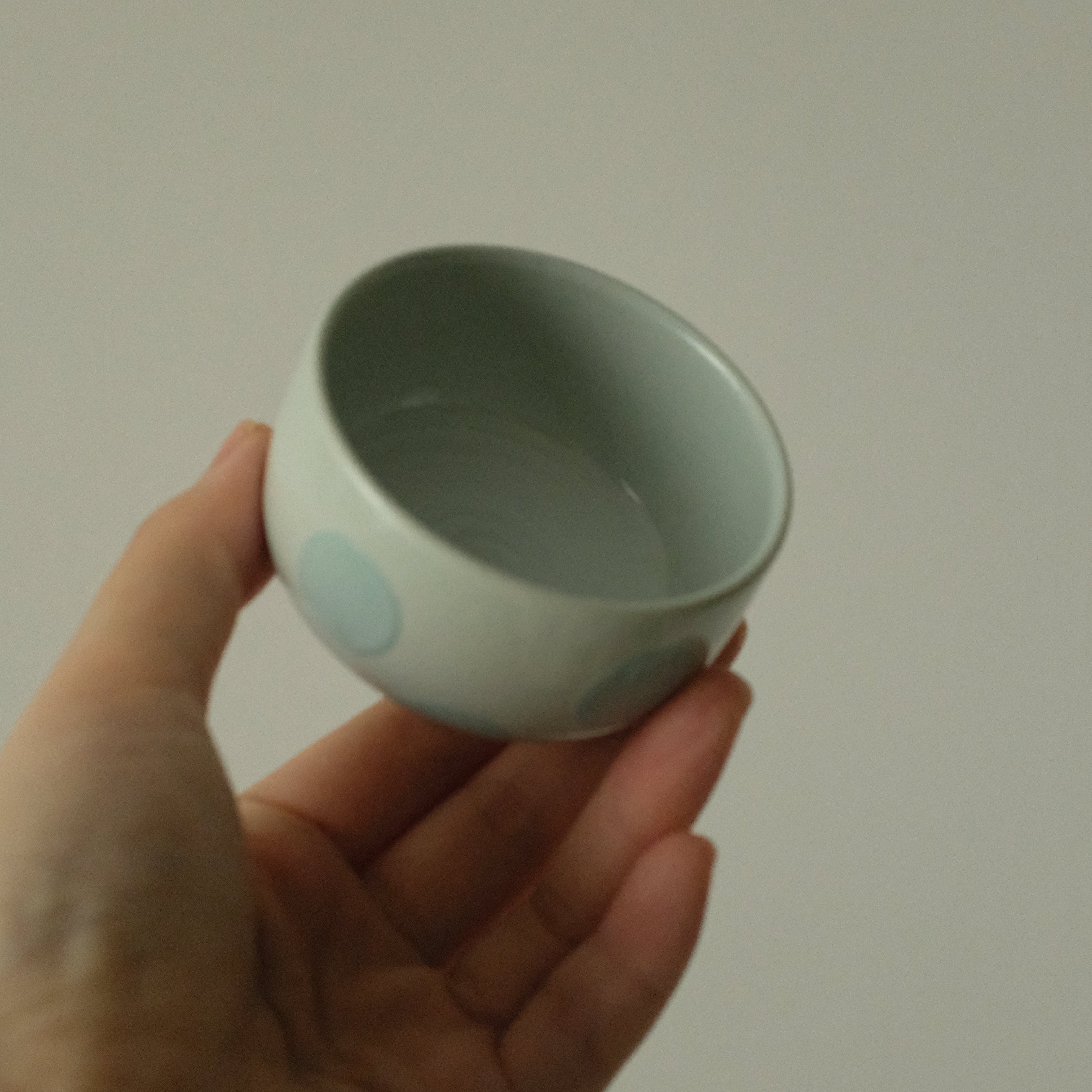 A hand holding a small ceramic cup with a pale blue-green glaze and light blue circular dot patterns, set against a plain light gray background.