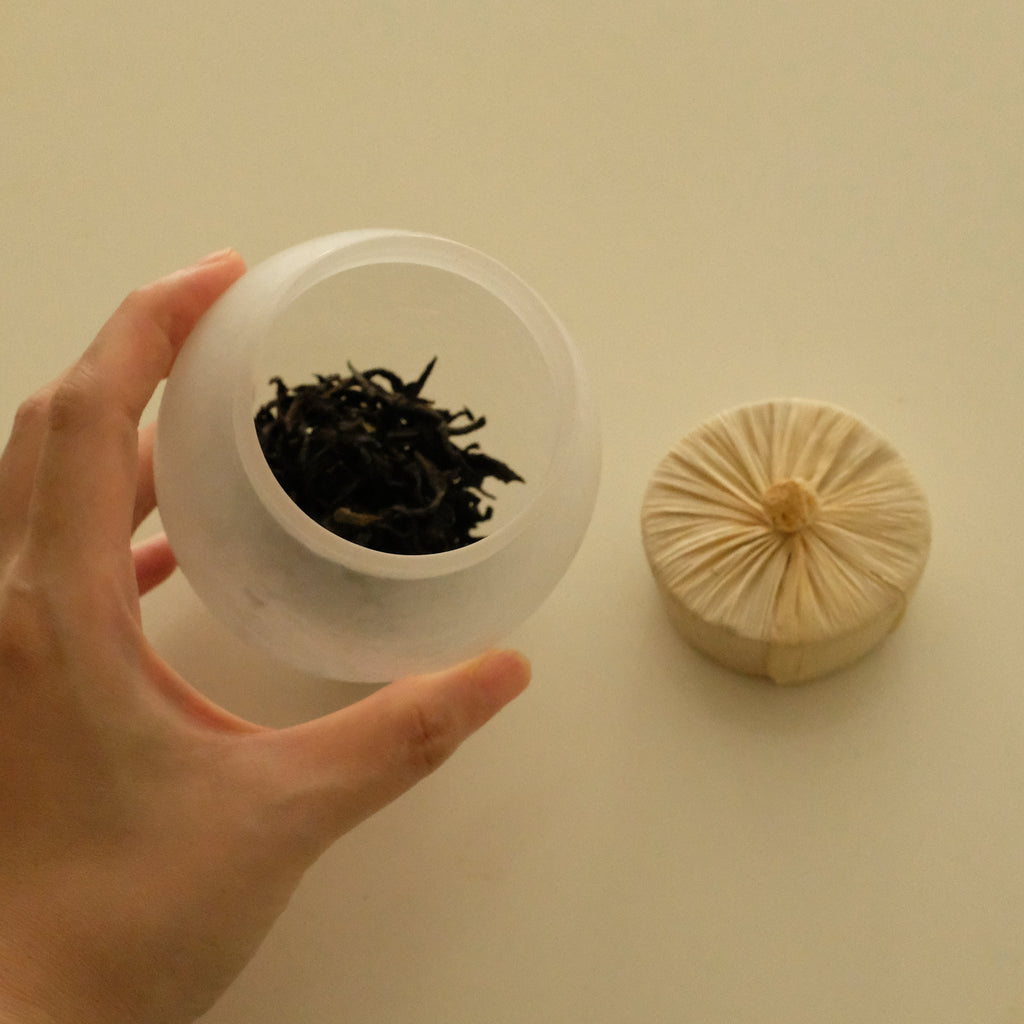 A hand holds a frosted clear plastic container filled with loose-leaf tea, with a round beige fabric lid placed beside it on a plain light background.