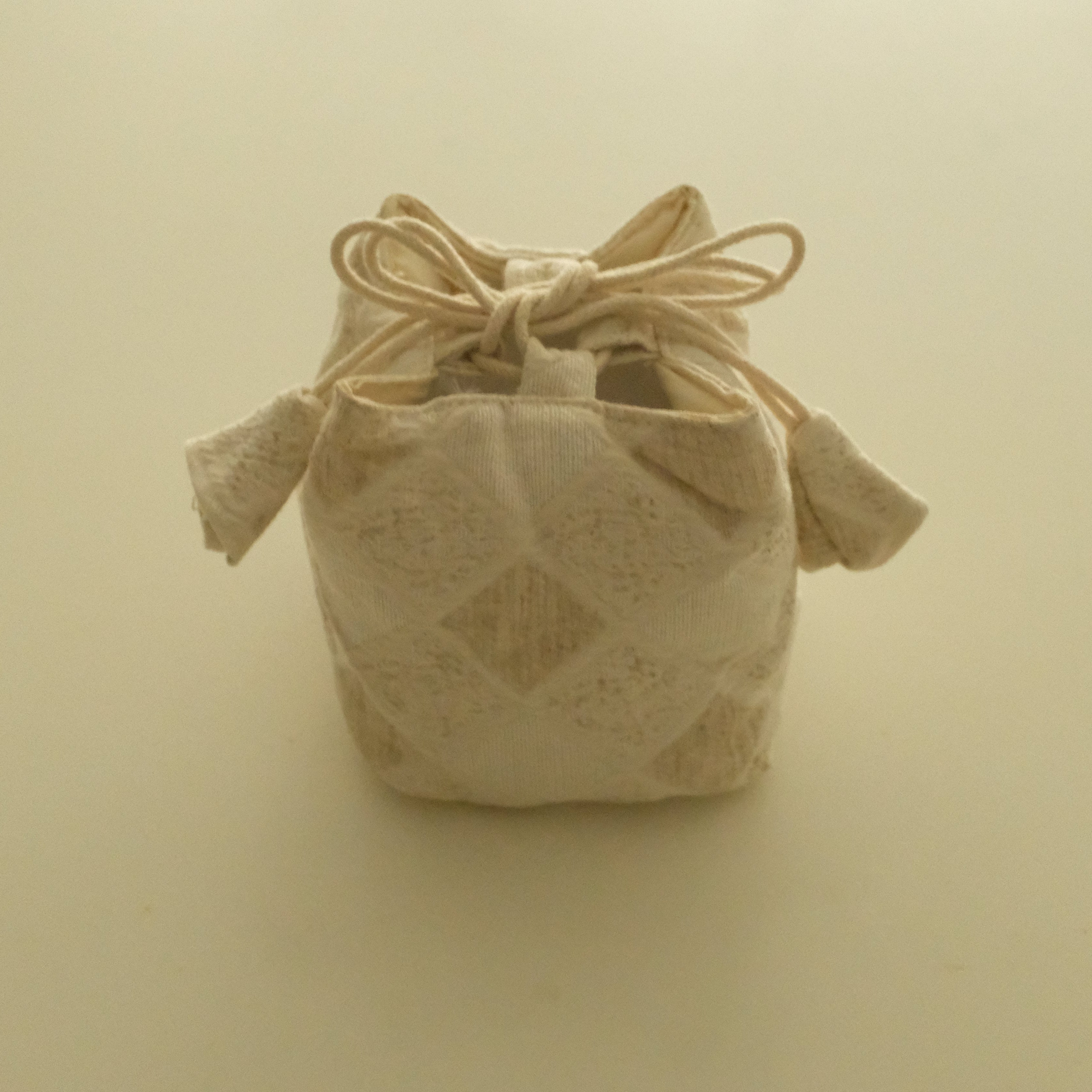 A small cream-colored fabric drawstring pouch with a subtle diamond pattern, photographed against a solid, warm off-white background.