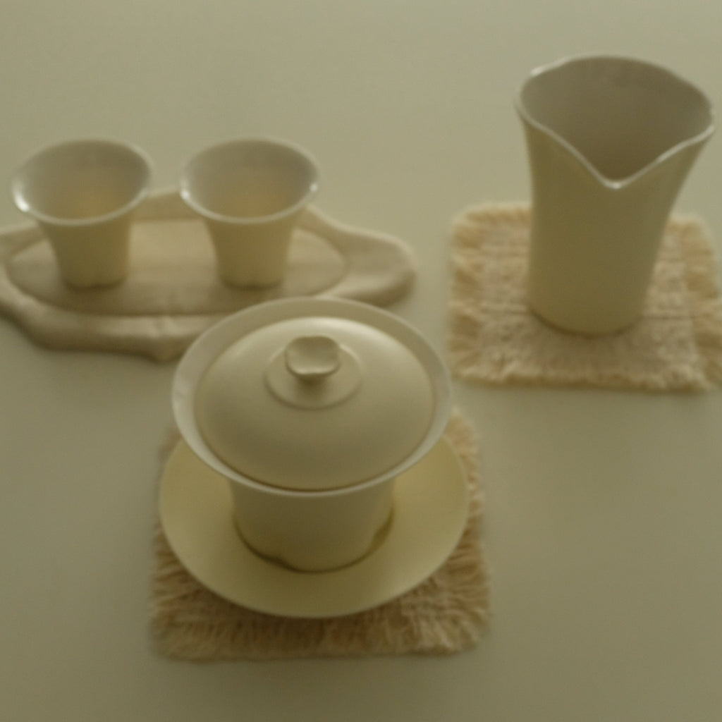 A set of cream-colored ceramic tea or coffee accessories, including a lidded teapot, two small cups on a tray, and a pitcher, arranged on textured beige cloth coasters against a solid cream background.
