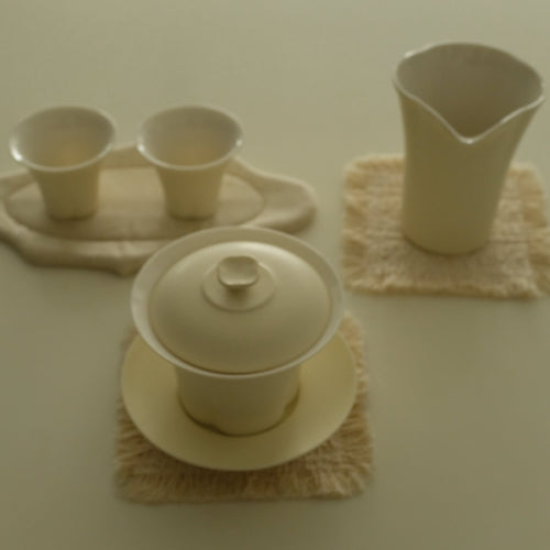 A set of cream-colored ceramic tea or coffee accessories, including a lidded teapot, two small cups on a tray, and a pitcher, arranged on textured beige cloth coasters against a solid cream background.