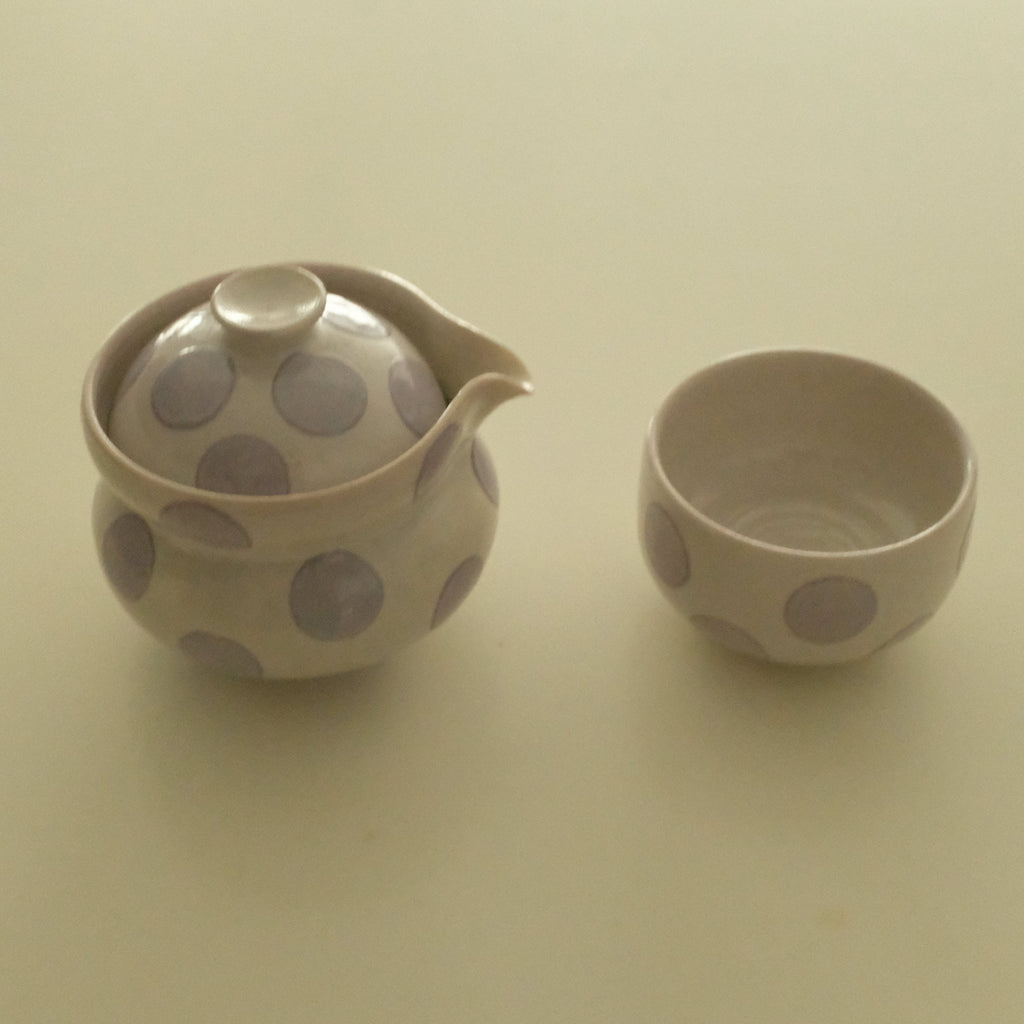 A small ceramic handless teapot and matching cup with a beige finish and lavender polka dots, set against a plain beige background.
