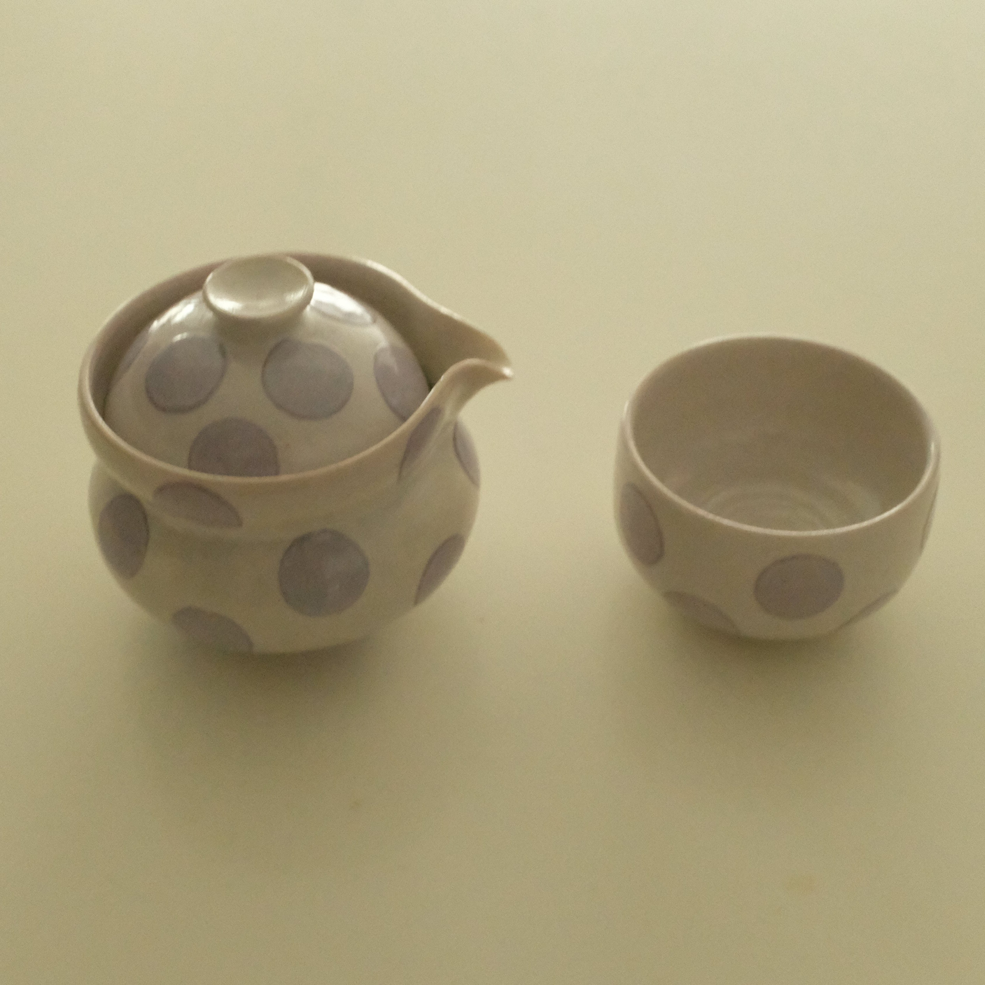 A small ceramic handless teapot and matching cup with a beige finish and lavender polka dots, set against a plain beige background.