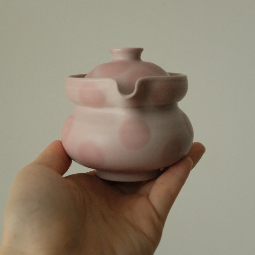 A hand holding a small pink ceramic teapot with a matching lid against a plain light gray background.