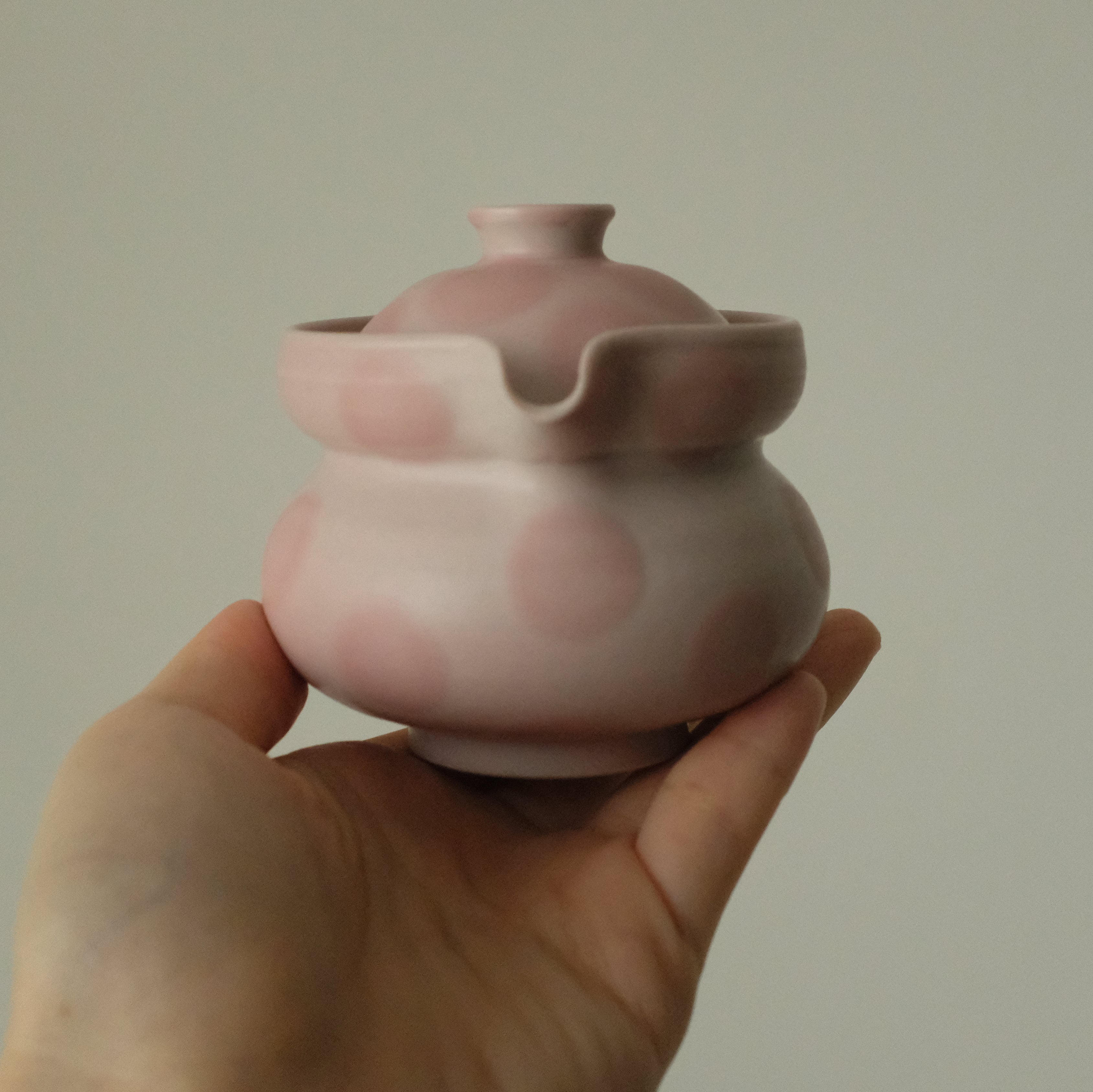 A hand holding a small pink ceramic teapot with a matching lid against a plain light gray background.