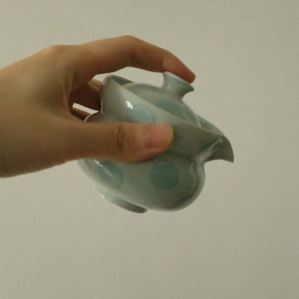 A hand holding a small glossy ceramic teapot with a pale blue-green glaze and circular dot patterns, set against a plain light gray background.