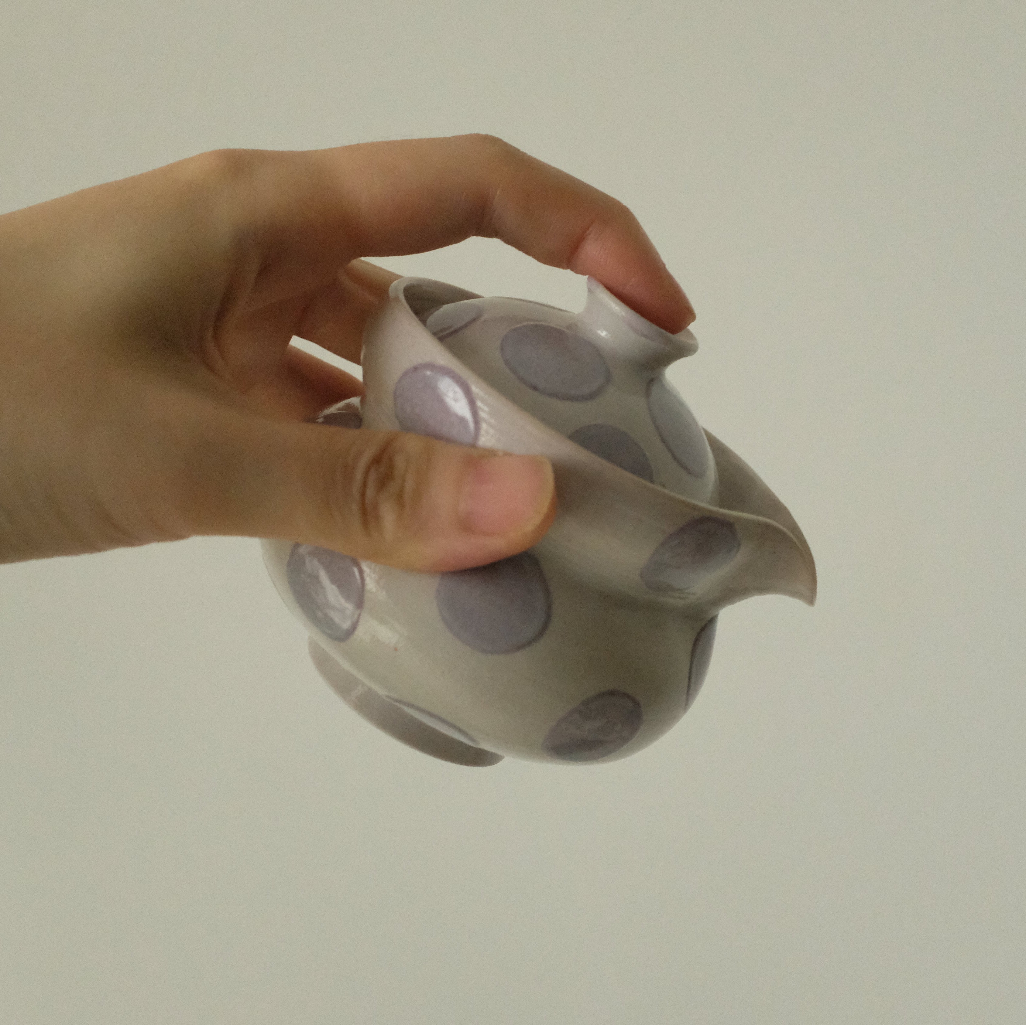 A hand holding a small white ceramic teapot with gray circular patterns, set against a plain light gray background.