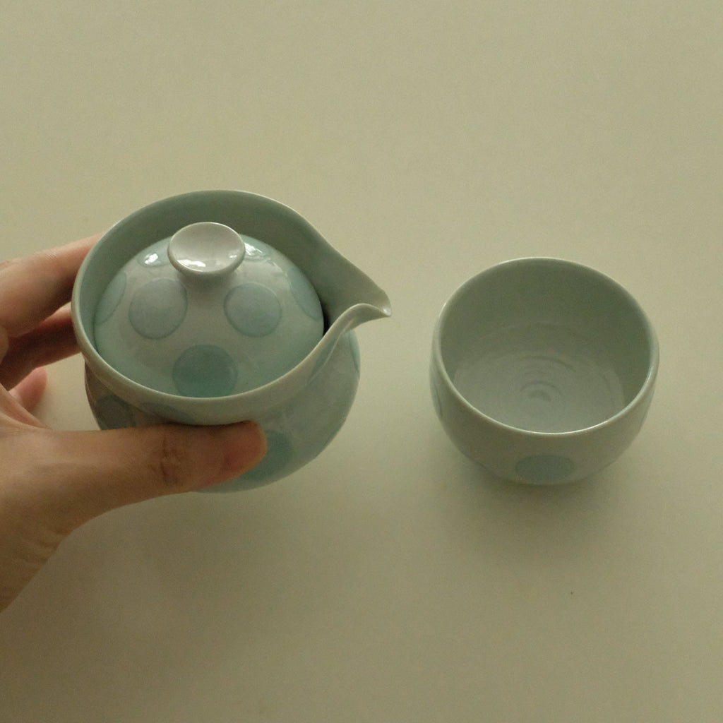 A hand holds a pale green ceramic teapot with a matching lid and a separate matching cup on a plain light beige background.