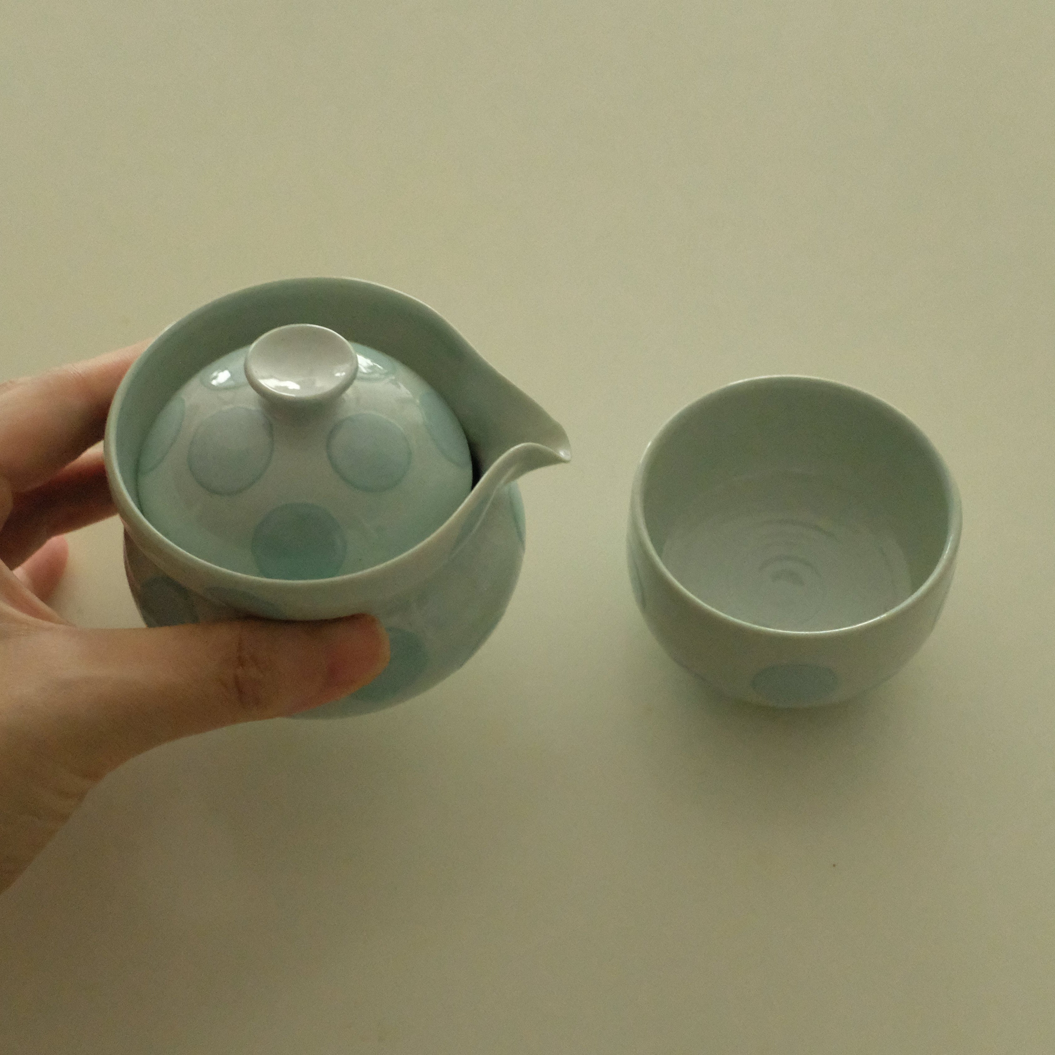 A hand holds a pale green ceramic teapot with a matching lid and a separate matching cup on a plain light beige background.
